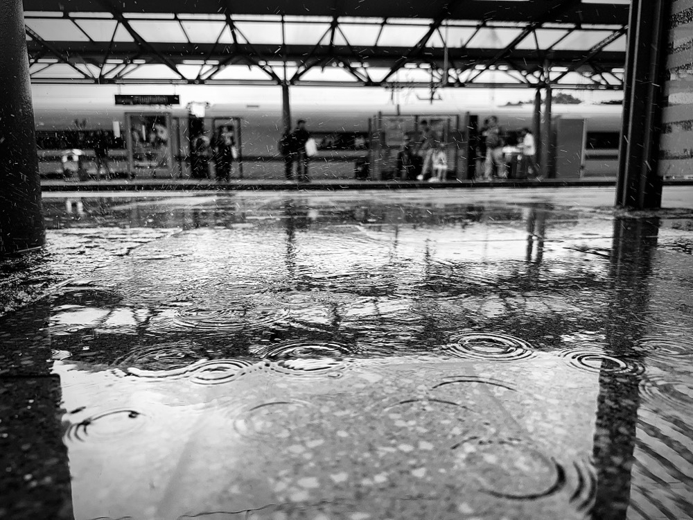 Black and white photo of water drops in the rain on a train platform with passengers in the background waiting on their train. 