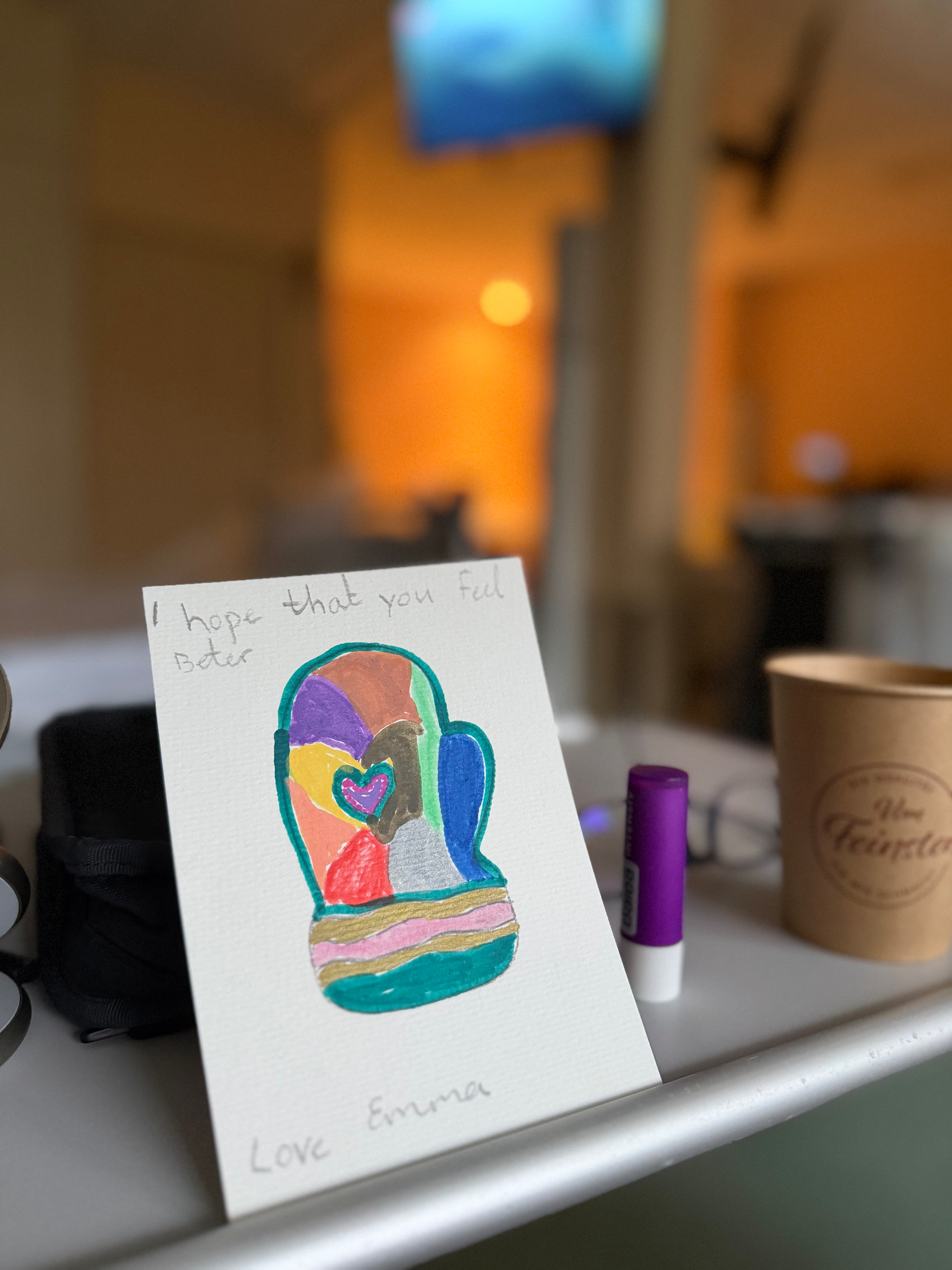 Hand drawn card propped up on a table next to a hospital bed. The card has a hand drawn mitten, with a heart in the middle of it, and the mitten is vibrantly colored with many colors. The card reads, “I hope that you feel better, love Emma”