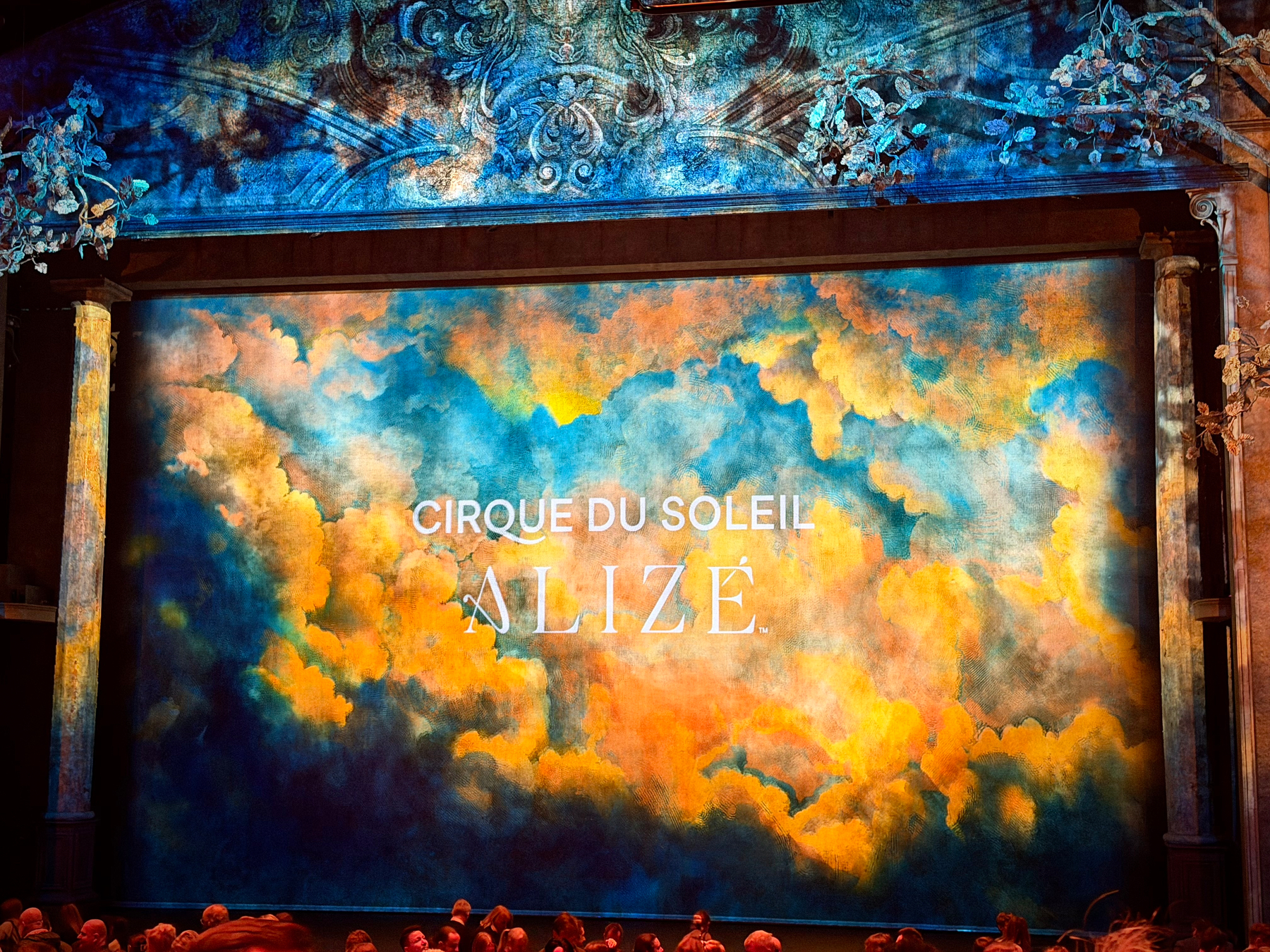 Theater stage showing a banner with clouds in blues, yellows, and oranges. Similar to a sunset. The text Cirque Du Soleil Alize is printed in the center of the banner over the clouds. 