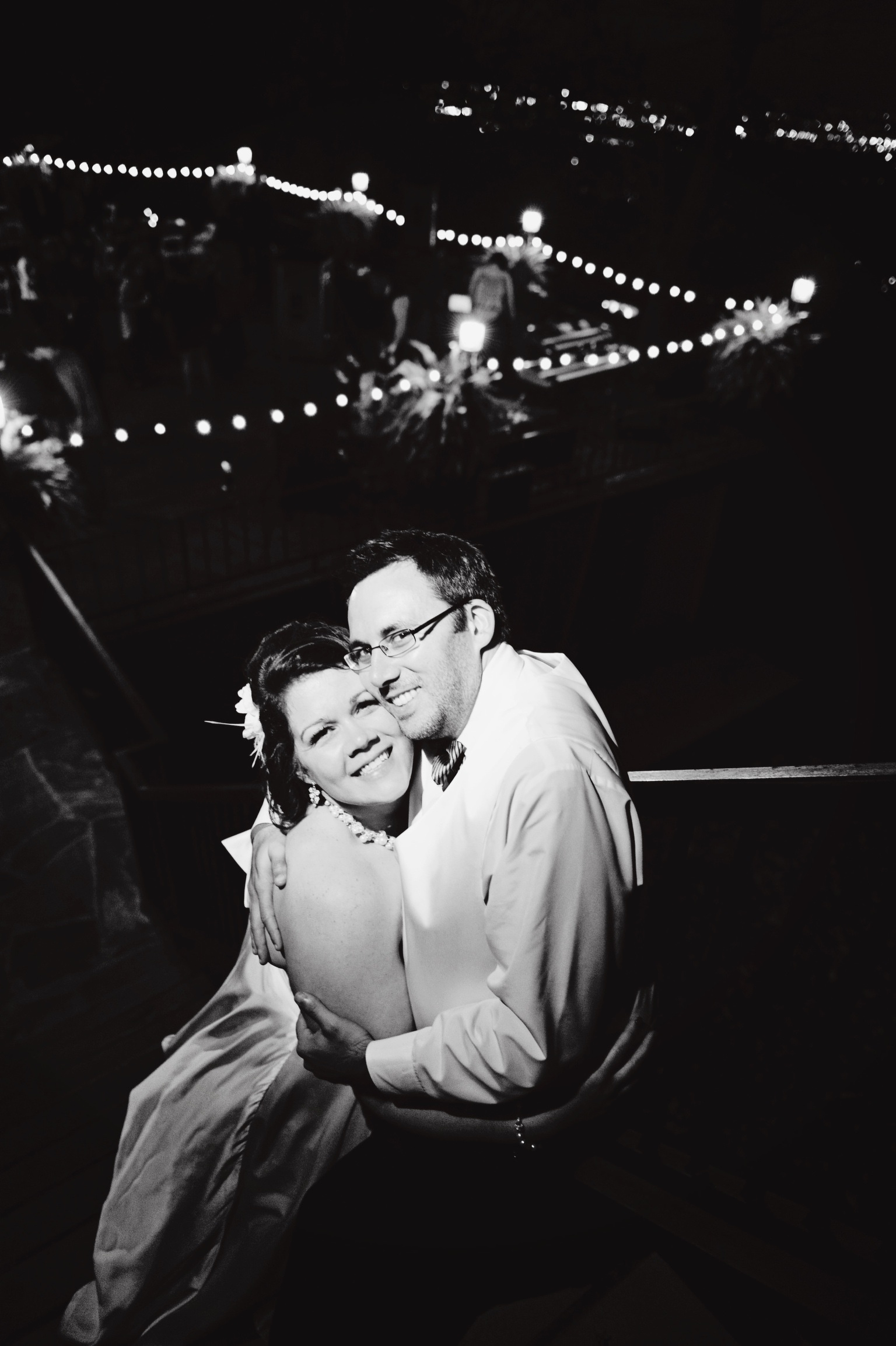Black and white photo of me hugging my wife, both looking into the camera, in the foreground. Blurred image of a terrace with a string of lights wrapping around the edge can be seen in the background.