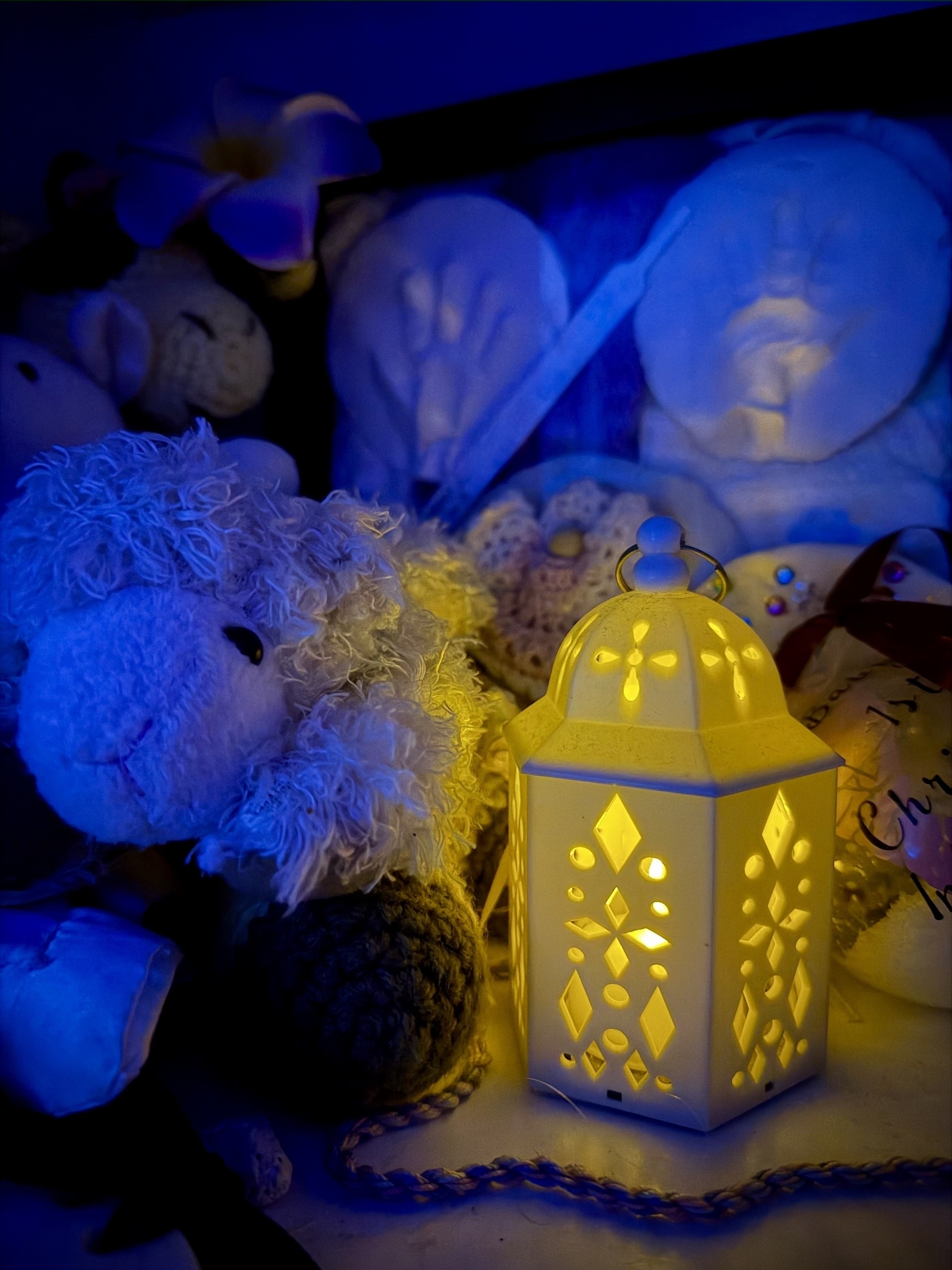Small candle inside of a case with shapes cut in the side to let light through. To the left of the candle is a stuffed lamb, with a stuffed giraffe behind it. Behind the candle to the right is a frame of some blankets and handprint molds of a baby.