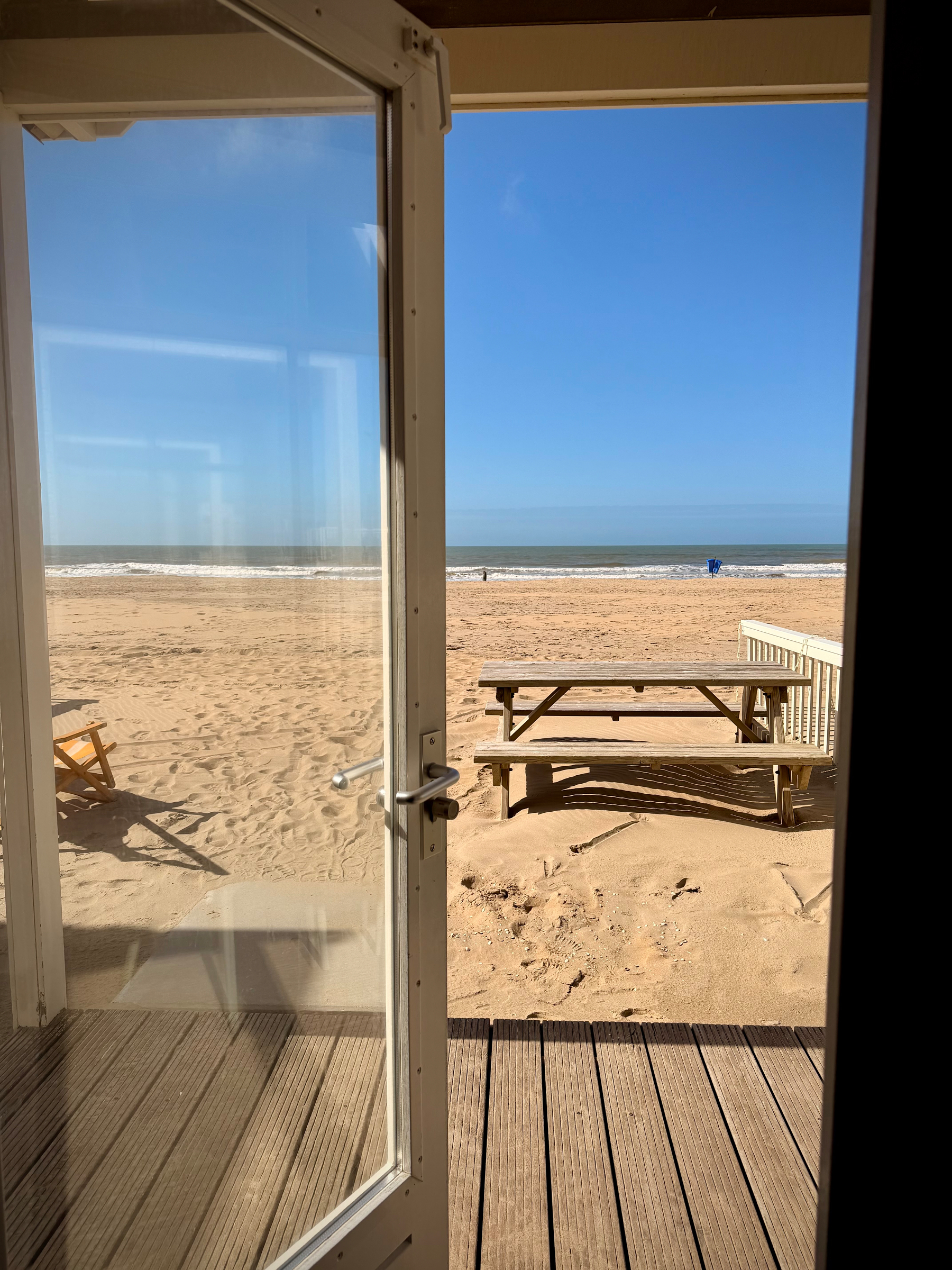 View out of a partially opened glass door onto the beach. A picnic table is sitting in the sand directly in front of the beach house.