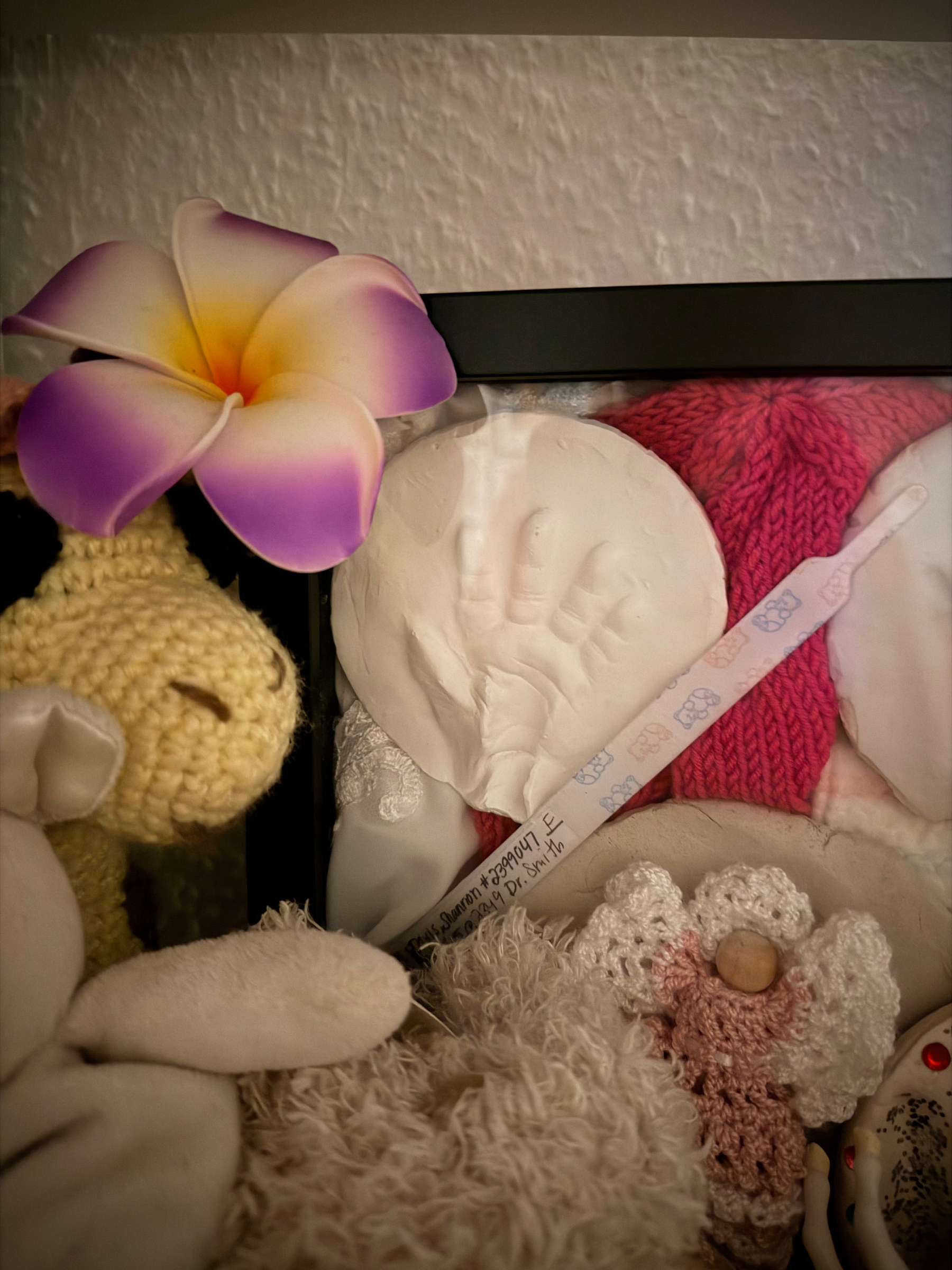 Framed mold of a baby’s hand, with a hat and hospital band. A flower sitting on the top left of the frame, a small knitted giraffe to the left of the frame and another small stuffed animal  at the bottom. 