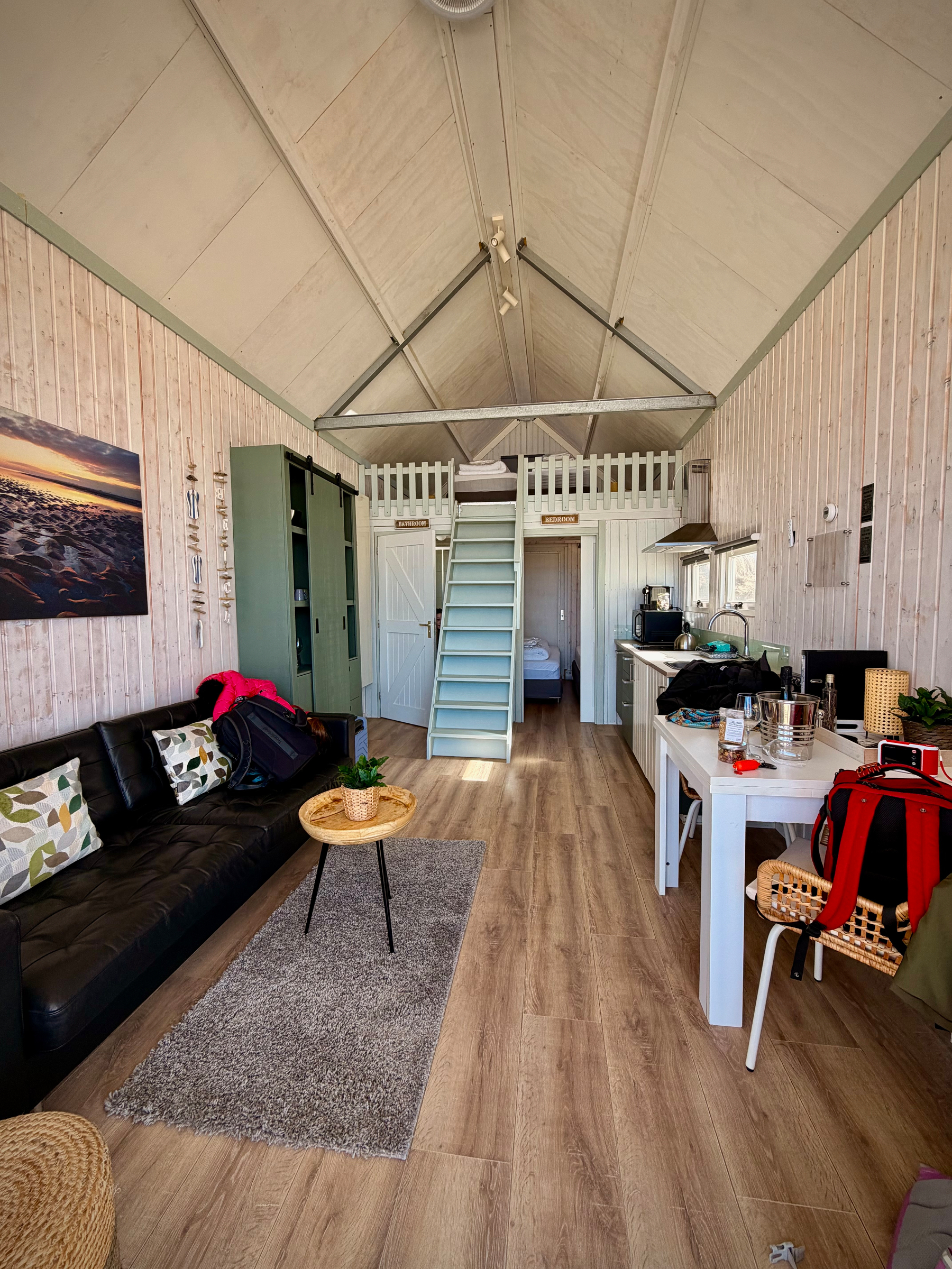Picture entering a little beach house, with a couch to the left, stairs straight ahead leading to a loft, and two doors on either side of the stairs. A little table and kitchenette are on the right.