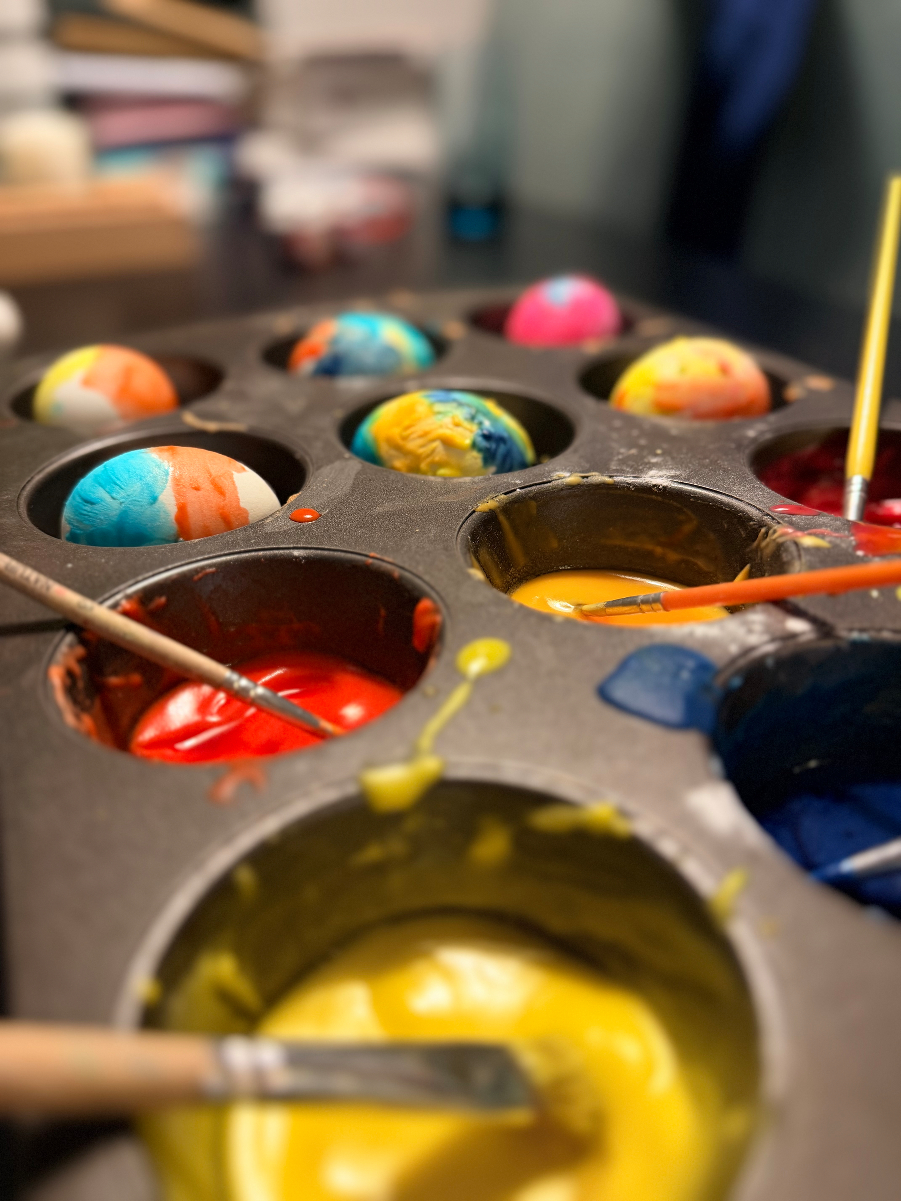 Cupcake pan with colors and paintbrushes in six of the slots and painted eggs in the other six. 