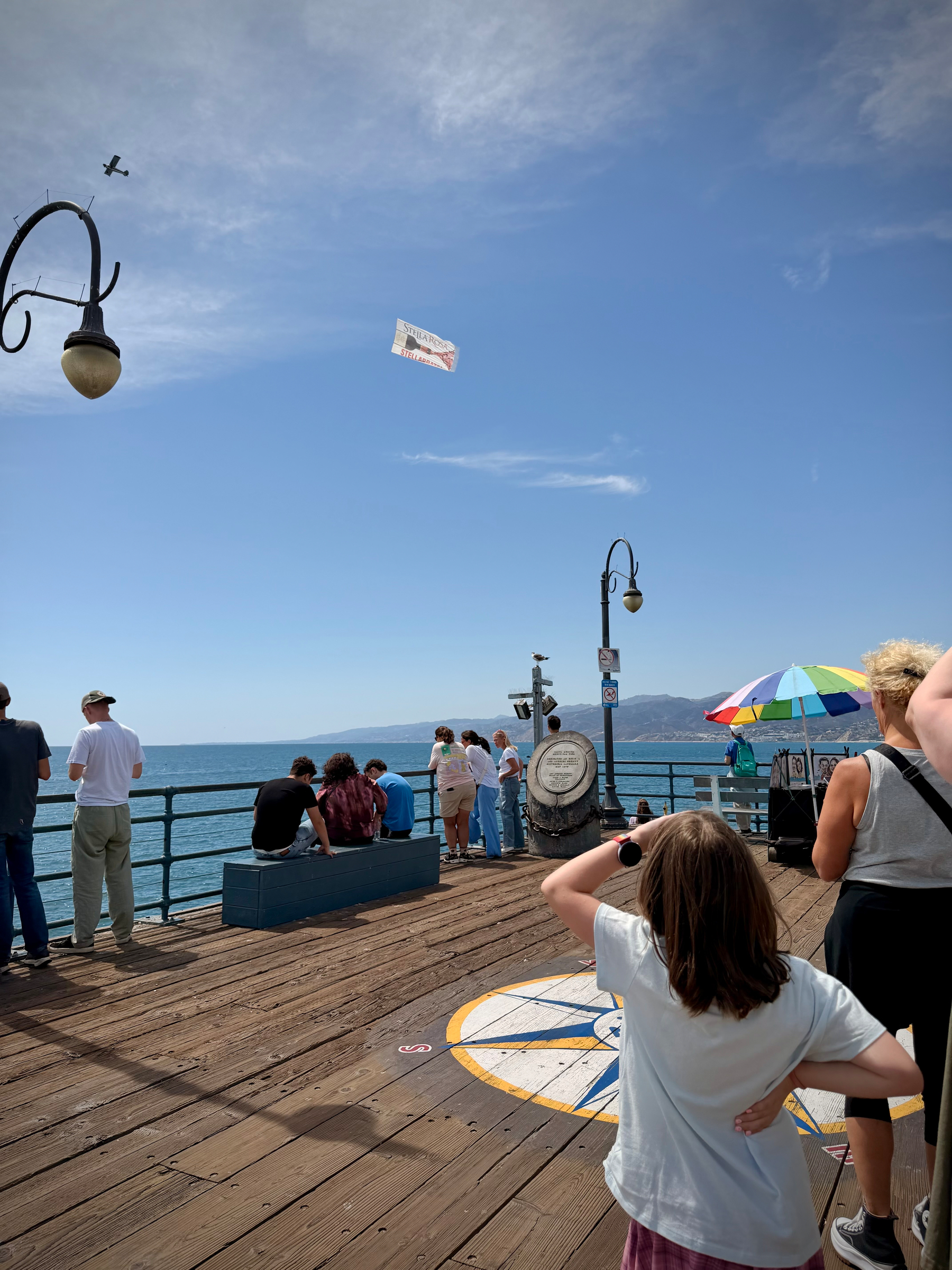 People standing at the end of the Santa Monica pier. A plane carrying a banner is flying overhead.