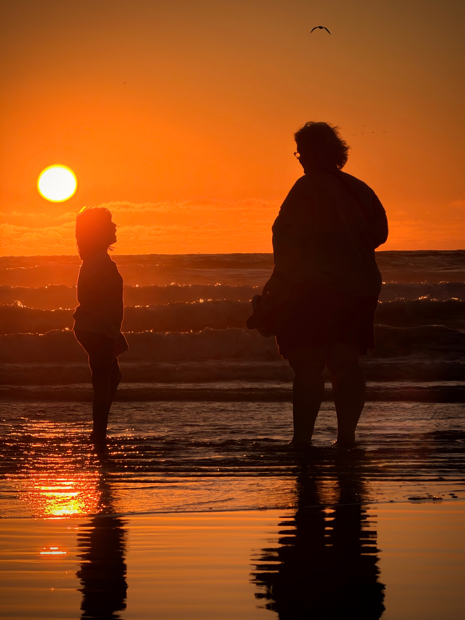 Sunset with the silhouette of a mom and daughter standing on the beach, slightly in the ocean.