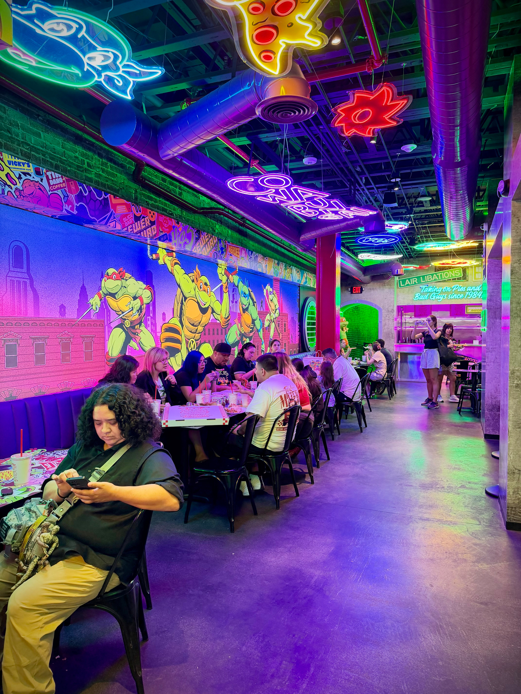 Dining area inside Teenage Mutant Ninja Turtles Pizzeria. Neon lights illuminating artwork of the turtles on the walls.