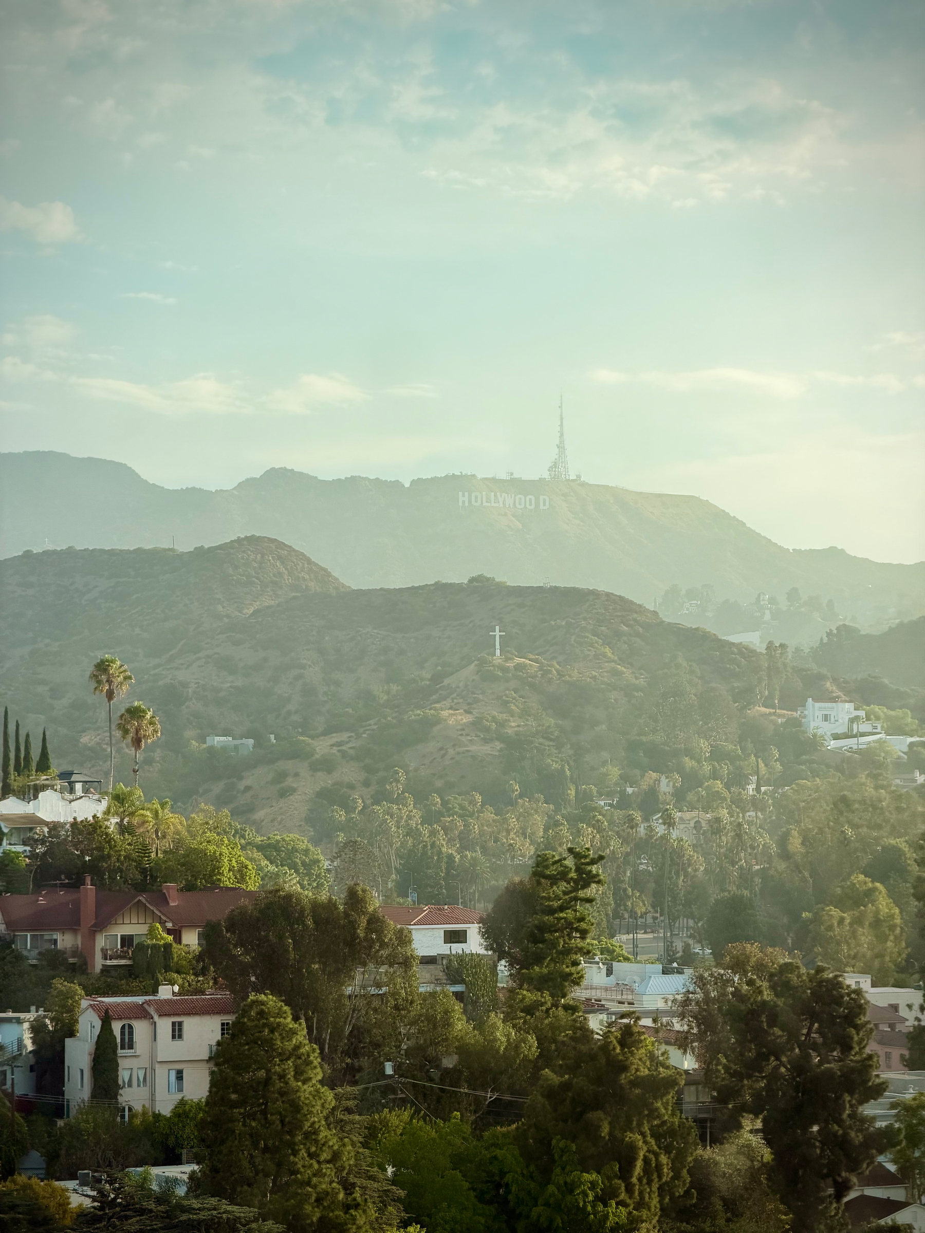 View of the Hollywood sign visible on the hills covered in a morning haze. Houses are visible through the trees in the foregr