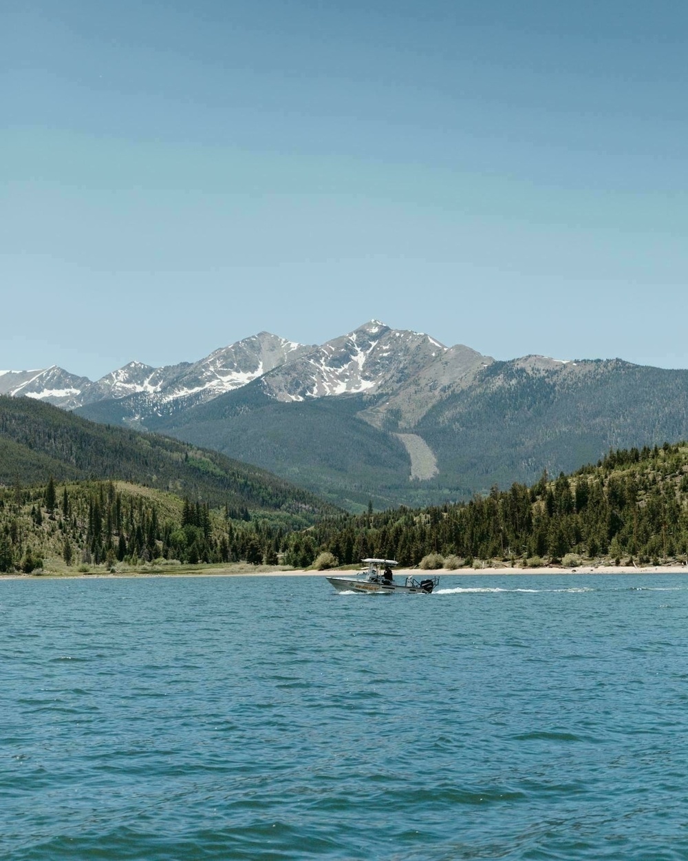 A speedboat glides across a lake surrounded by mountains and forested hills under a clear blue sky.