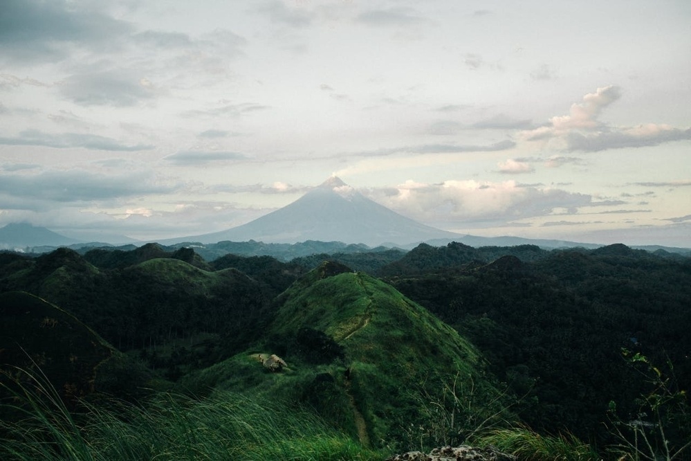 A picturesque landscape features rolling green hills with a distant view of a volcanic mountain under a cloudy sky.