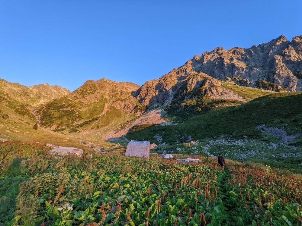 A small wooden cabin is nestled in a lush, green mountain valley under a clear blue sky.