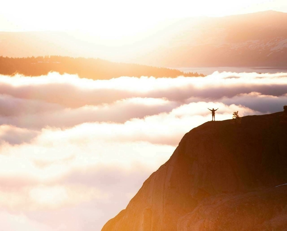 A person stands triumphantly on a cliff edge overlooking a breathtaking scene of clouds and a glowing sunset.