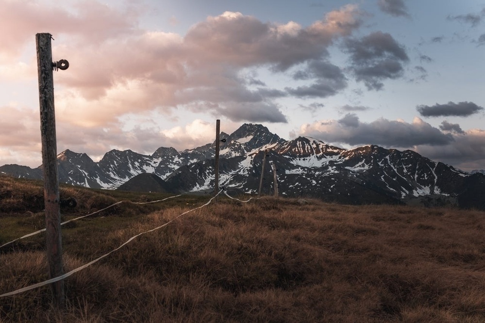 A mountain range with snow patches is seen behind a grassy foreground and a fence at sunset.