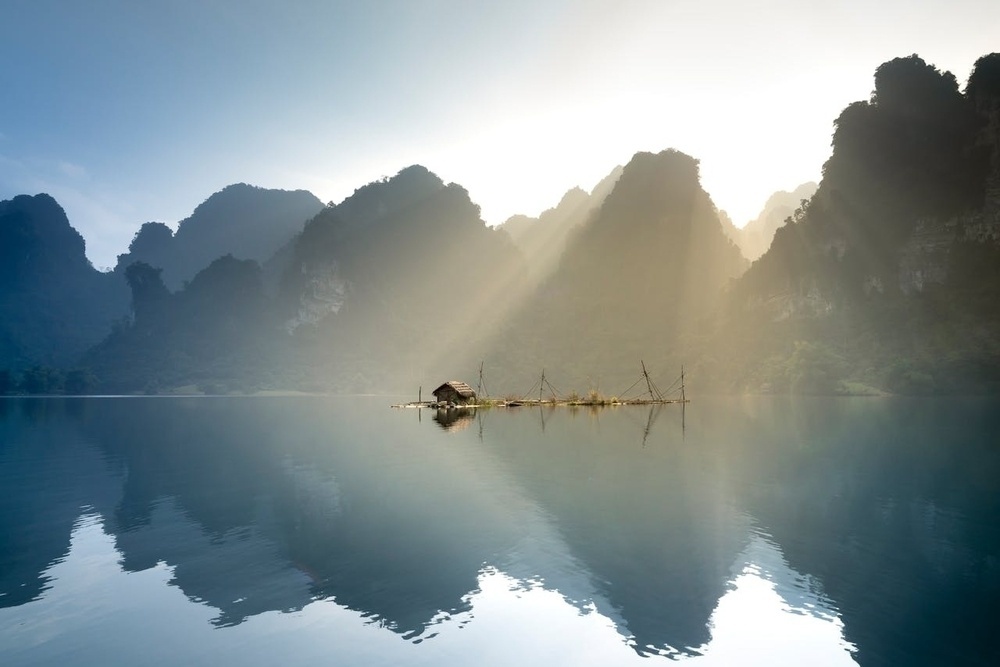 A serene lake reflects the silhouettes of distant mountains and a small hut as sunlight streams through the misty landscape.