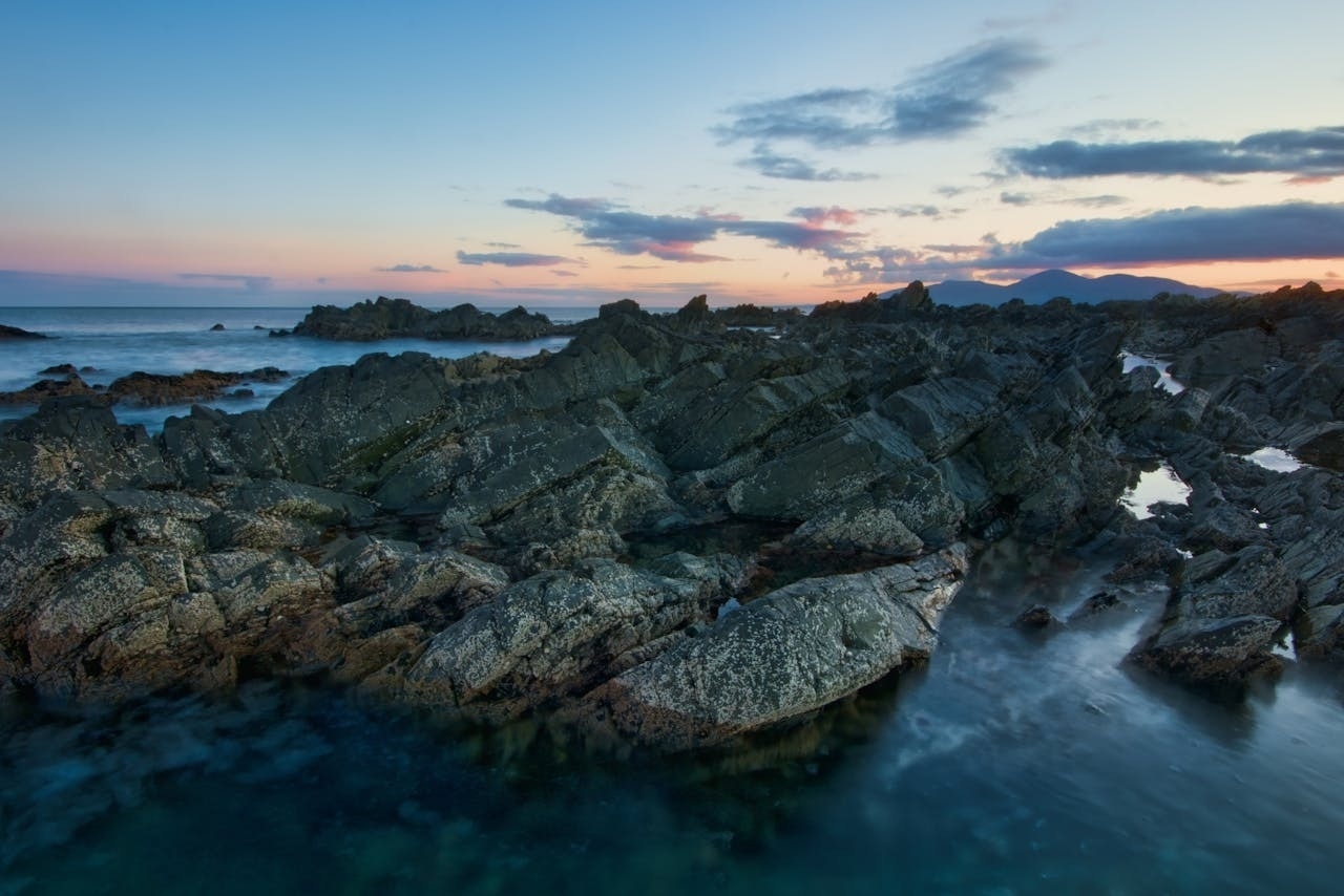 Auto-generated description: A rocky coastline is bathed in the soft light of sunset with calm ocean waters and a colorful sky.