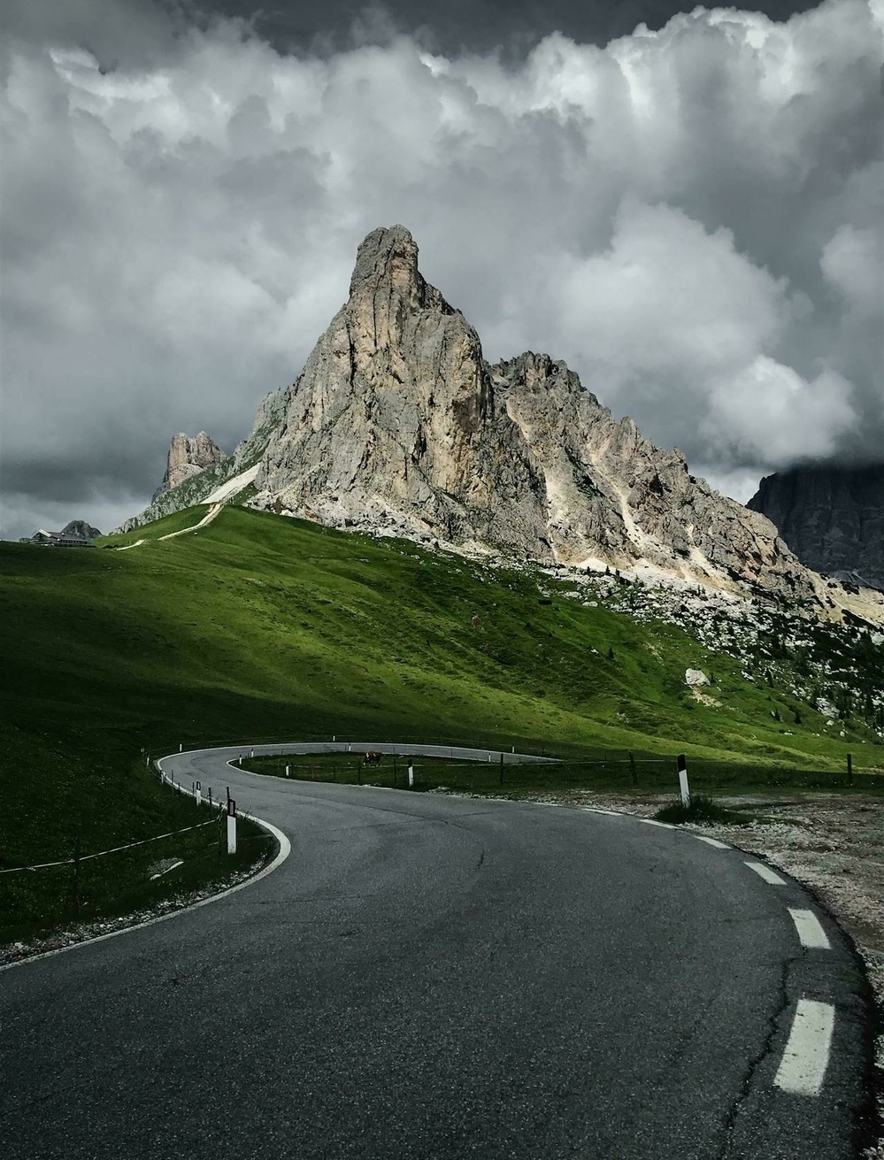 A winding road leads towards a jagged mountain under dramatic, cloudy skies.