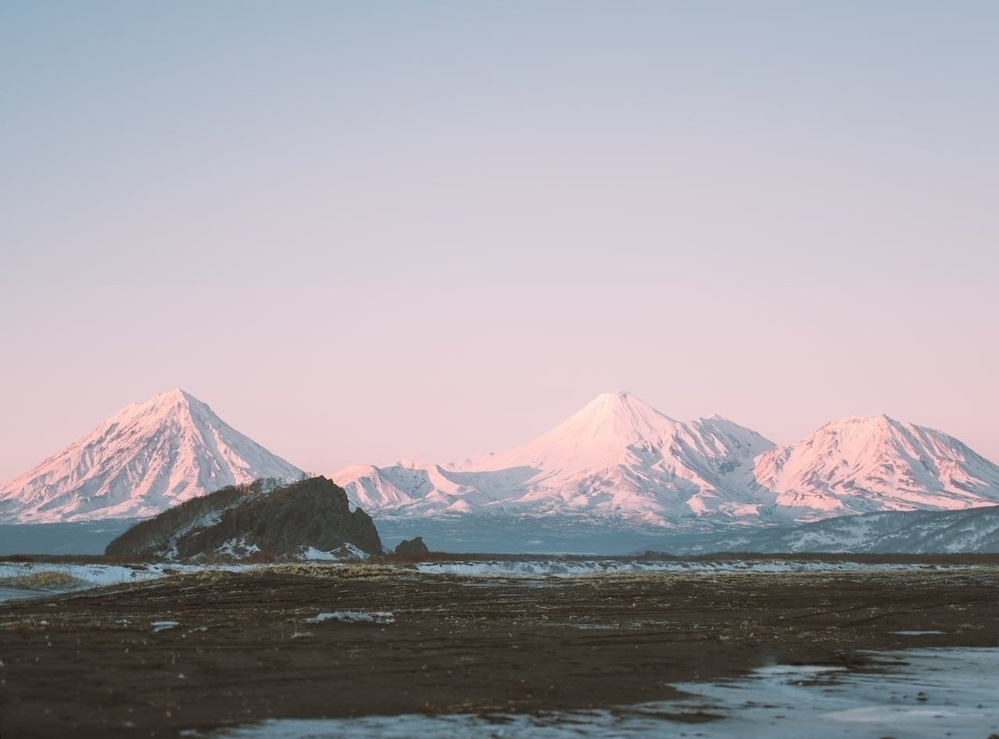 Snow-capped mountains are illuminated by soft pink light under a clear sky, with a barren, rocky foreground.