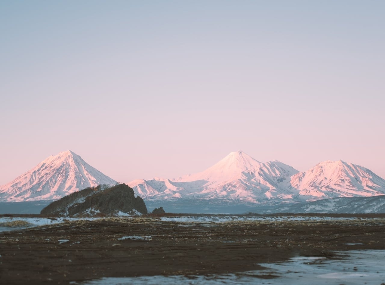 Auto-generated description: Snow-capped mountains are illuminated by soft pink light under a clear sky, with a barren, rocky foreground.