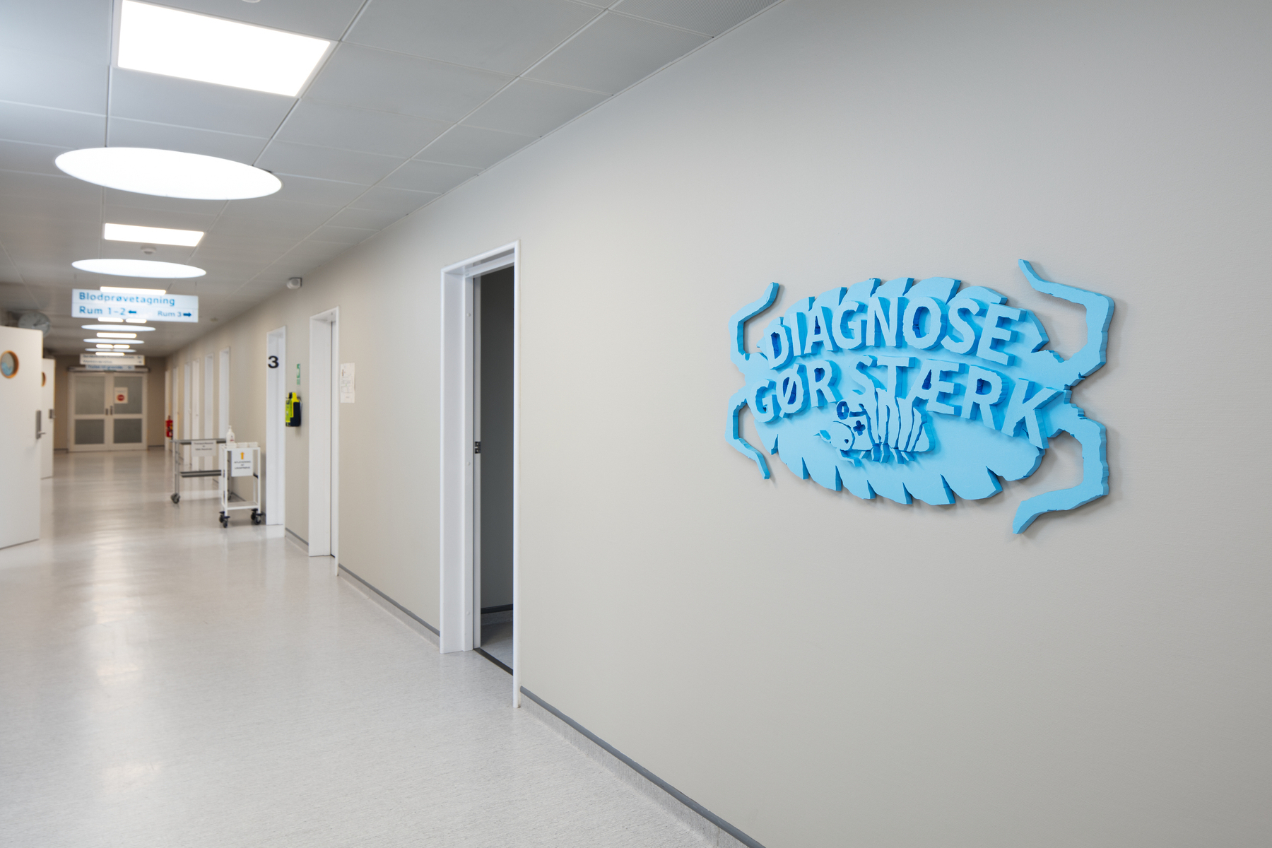 A hospital hallway features a light blue decorative sign on the wall with Danish text, alongside a corridor lined with doors and medical equipment.