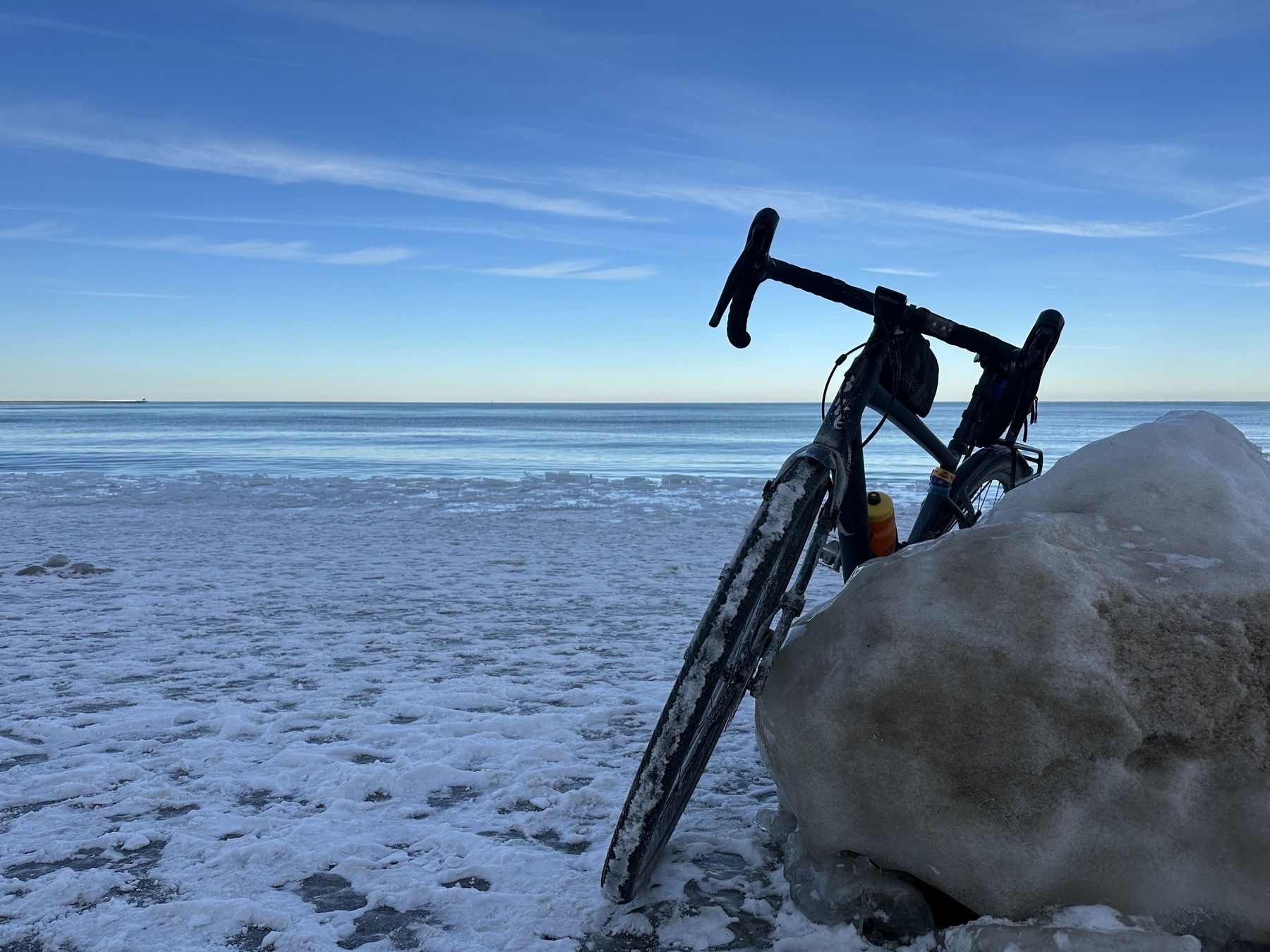 A bicycle rests against an ice boulder on a snowy lakefront with calm water and clear sky in the background.