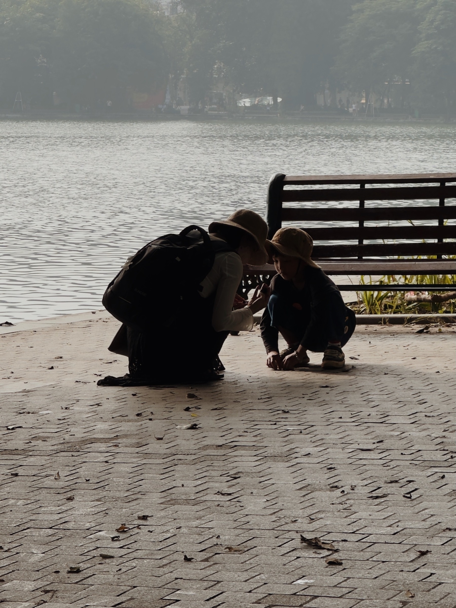 Auto-generated description: A person and a child wearing hats are kneeling and looking at something on the ground near a lakeside bench.