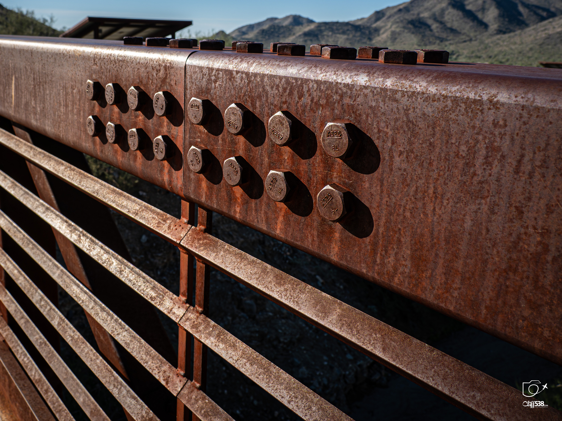Auto-generated description: A rusted metal bridge railing with large bolts is set against a mountainous landscape in the background.