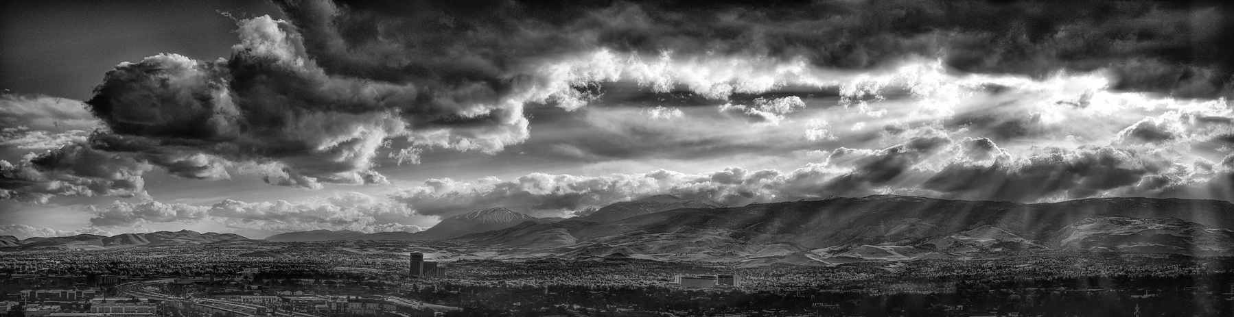 Auto-generated description: Dramatic clouds and sun rays dominate the sky over a sprawling cityscape with mountains in the background.