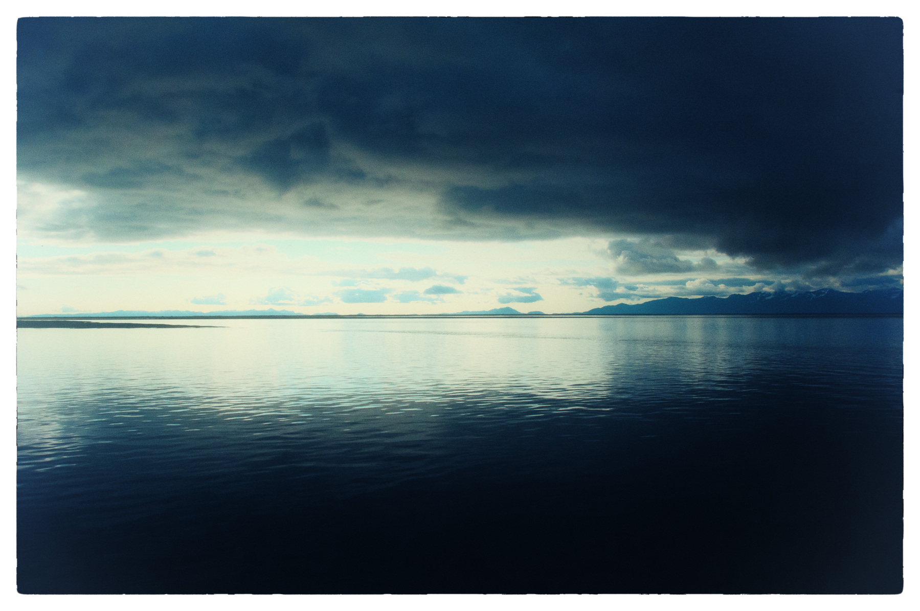 Auto-generated description: A vast, serene body of water reflects the moody clouds above, with a faint outline of distant land on the horizon.