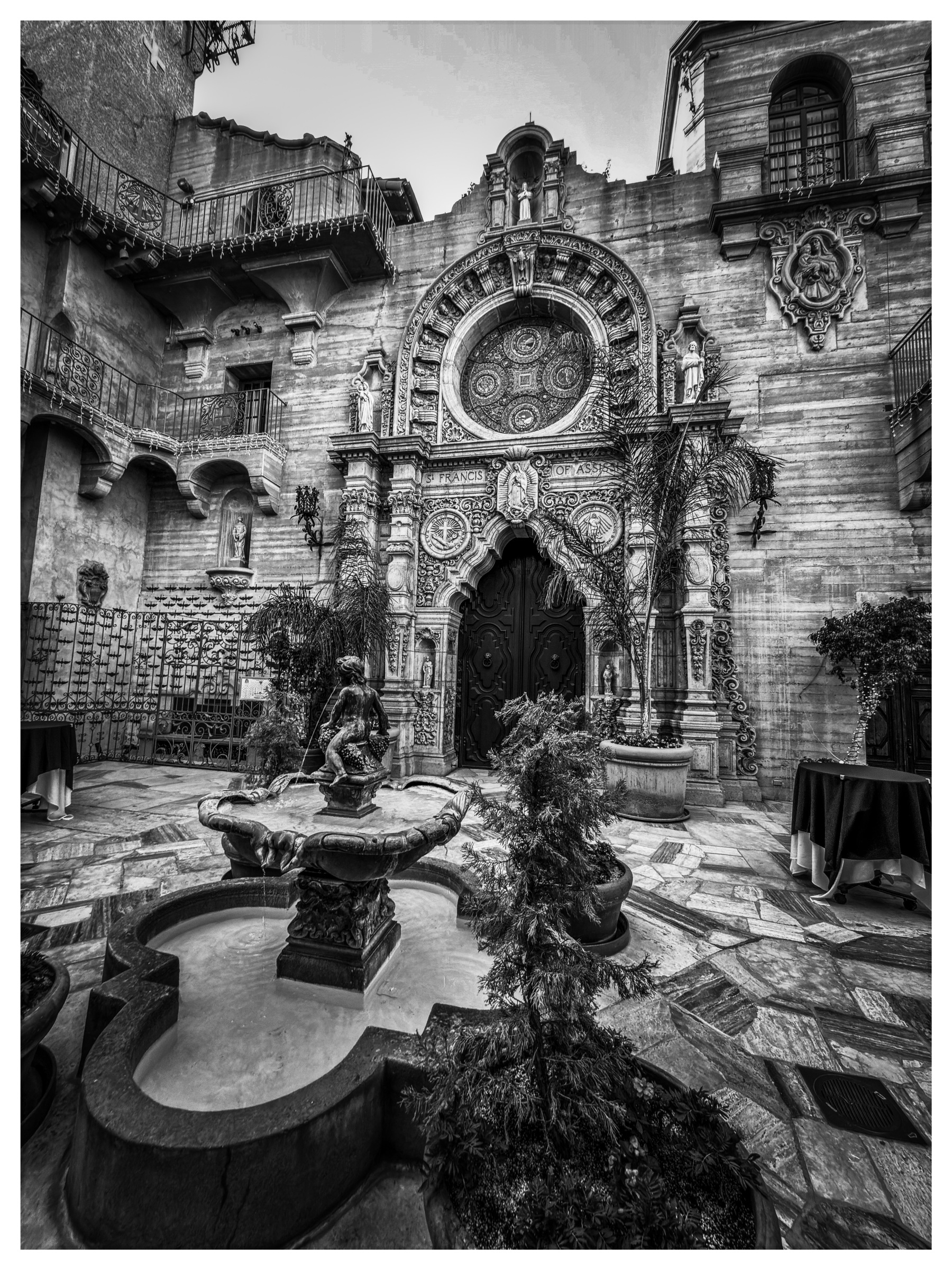 Auto-generated description: A historic, ornate courtyard with detailed stone architecture, a grand wooden door, and a central fountain surrounded by plants.