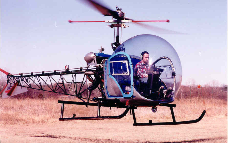 Auto-generated description: A person is flying a small, bubble-cockpit helicopter over a grassy field.