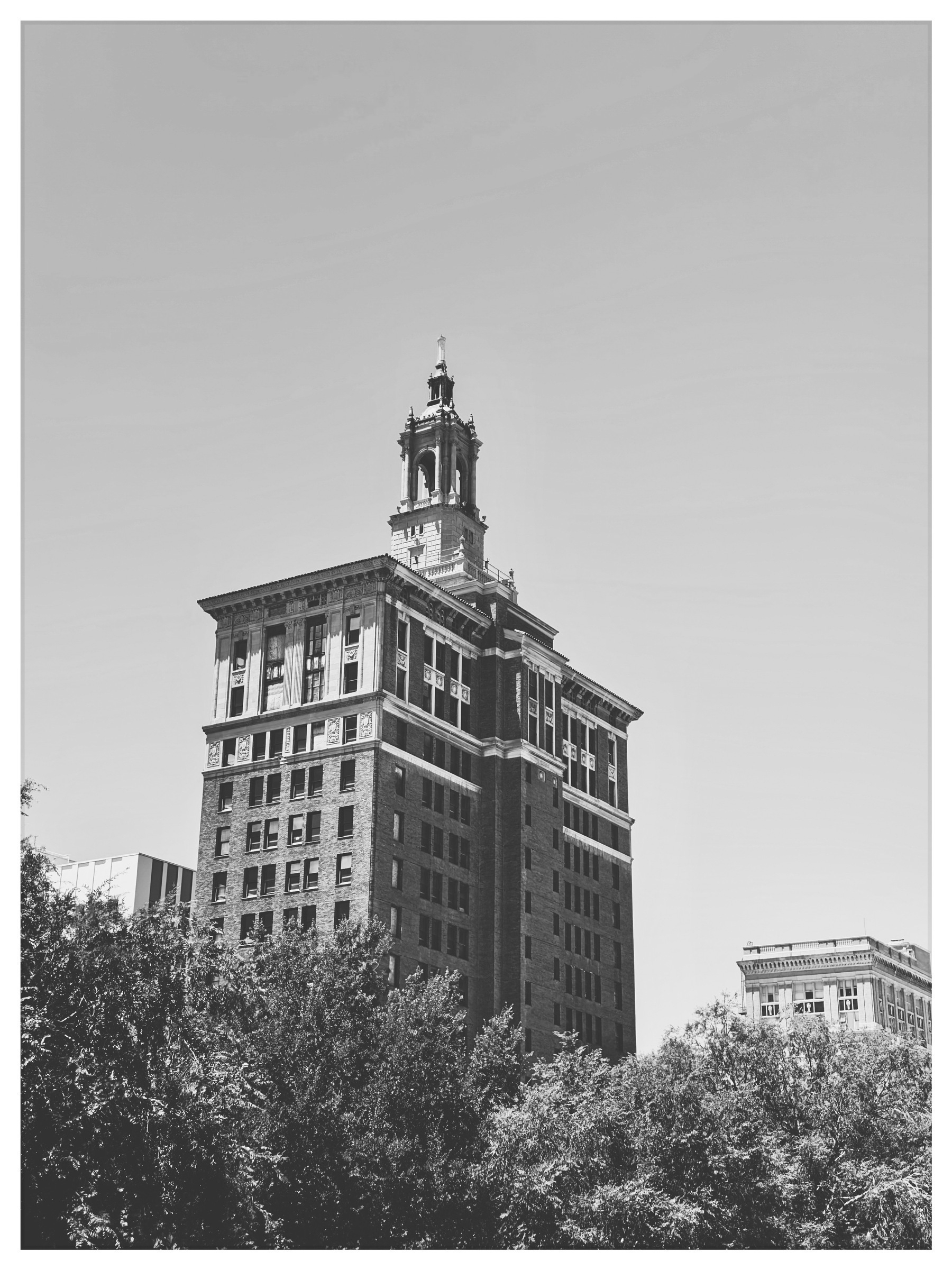 Auto-generated description: A historic multi-story brick building with a tall ornate tower rises above surrounding trees under a clear sky.