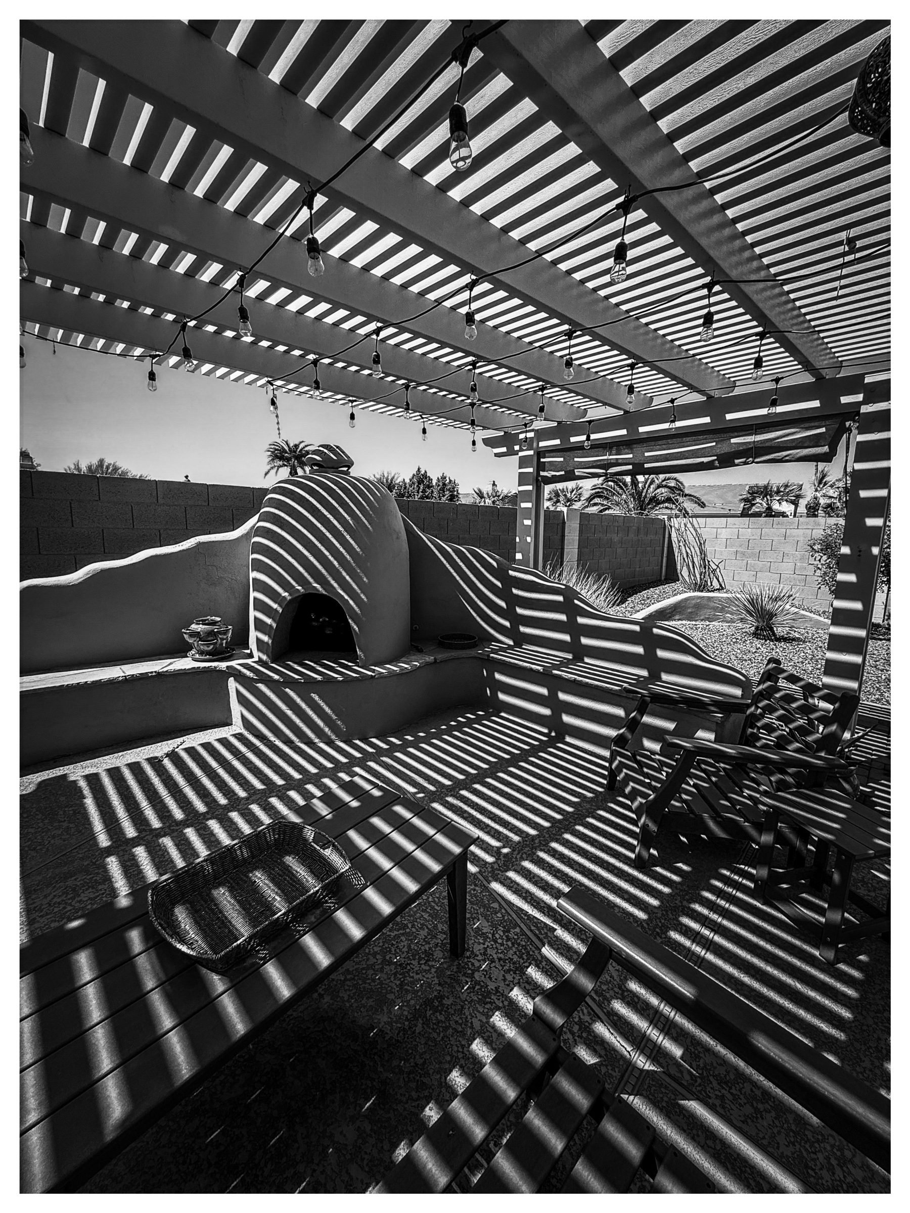 Auto-generated description: A sunlit patio features a pergola casting striped shadows, with lounge chairs and a fireplace.