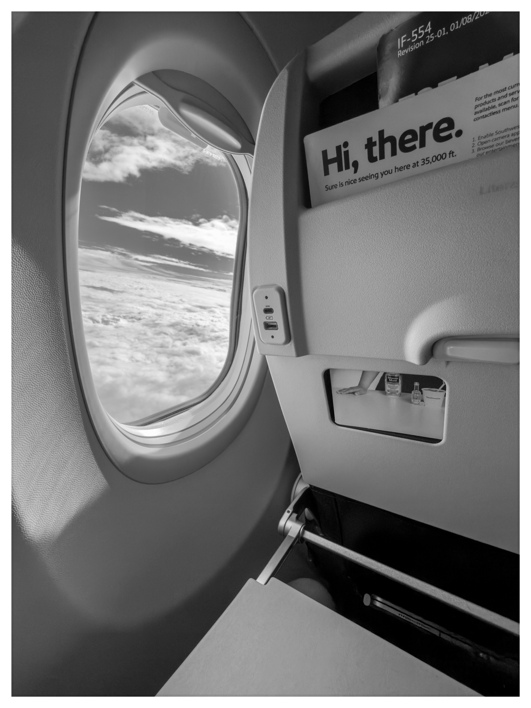 Auto-generated description: An airplane window seat view shows clouds and an open tray table, with a friendly greeting on the seat-back pocket.