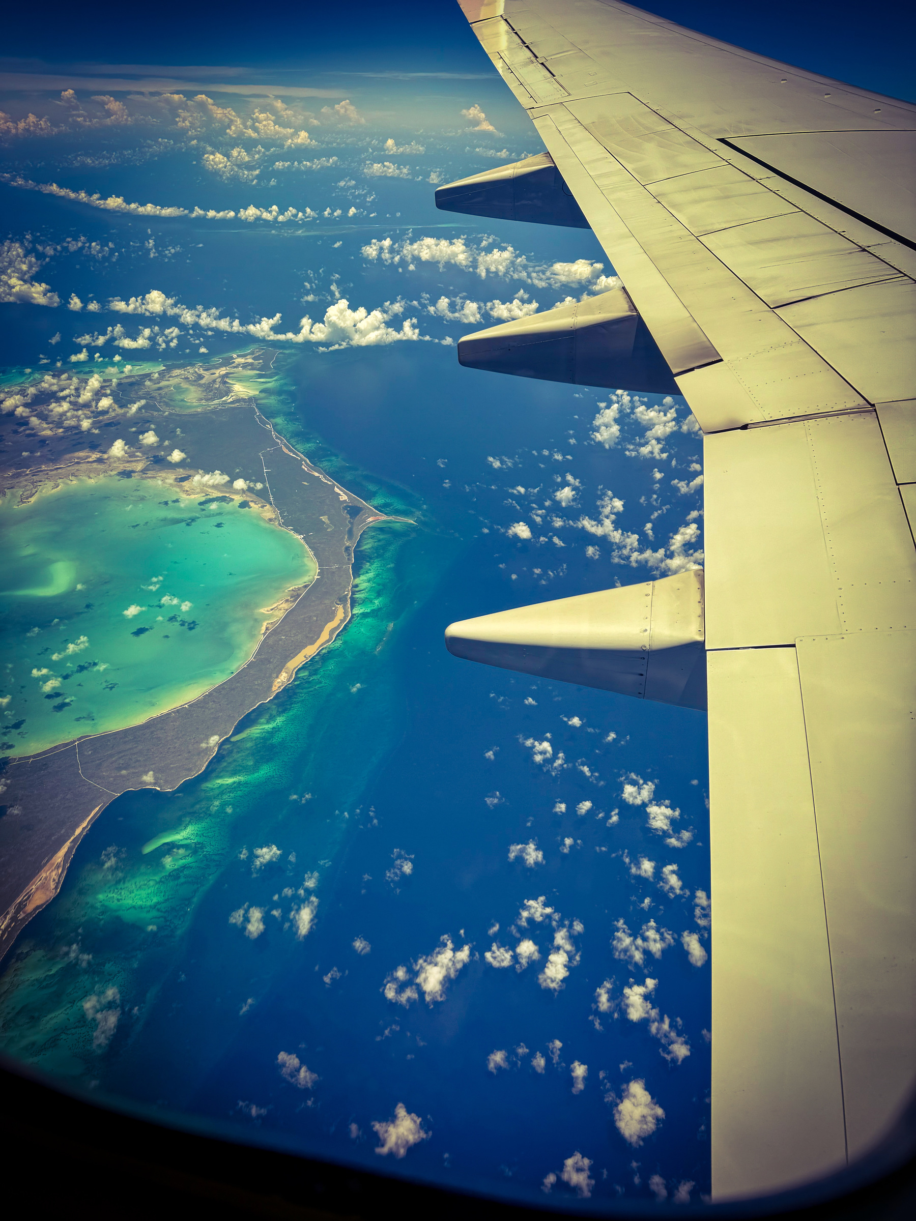 Auto-generated description: A view of a coastline and vibrant turquoise ocean is seen from an airplane window, with the aircraft's wing in the foreground.