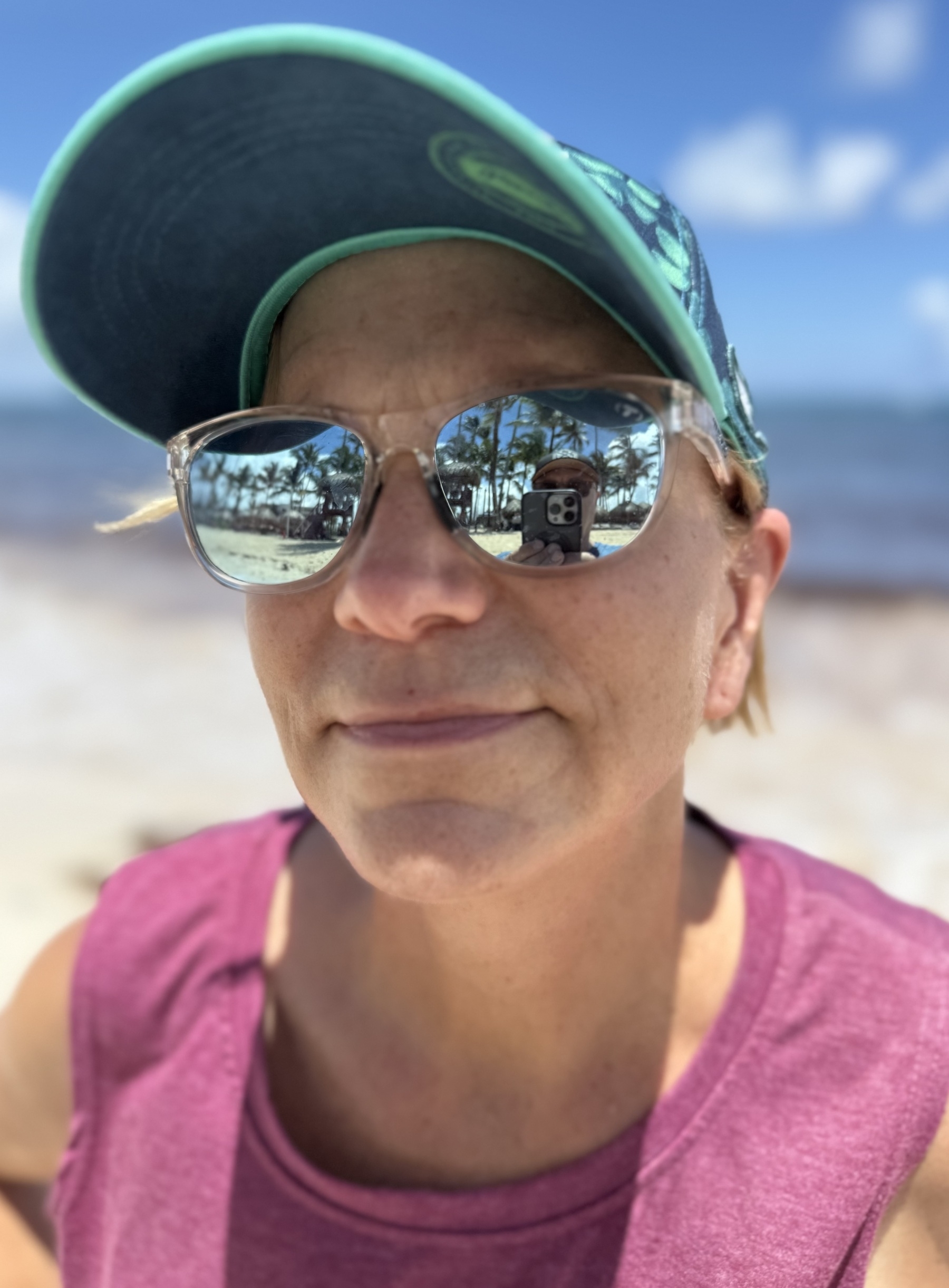 Auto-generated description: A person wearing sunglasses and a cap is on a sunny beach, with palm trees reflected in the lenses.