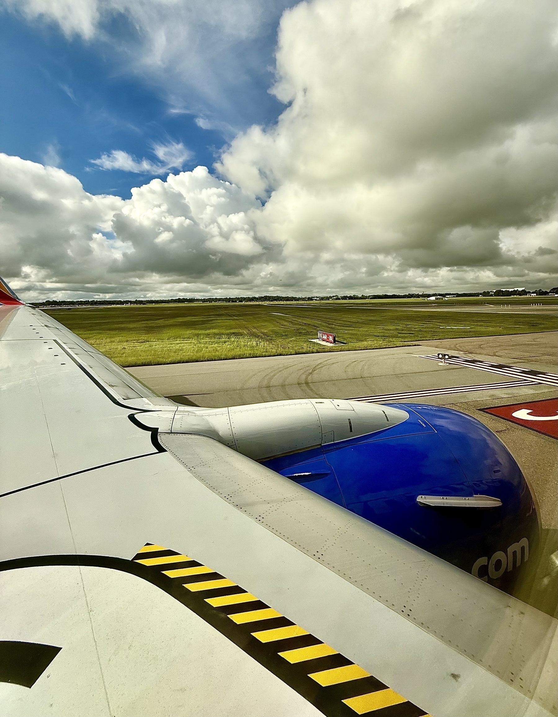 Auto-generated description: A view from an airplane window shows the wing and engine against a backdrop of a cloudy sky and grassy airfield.