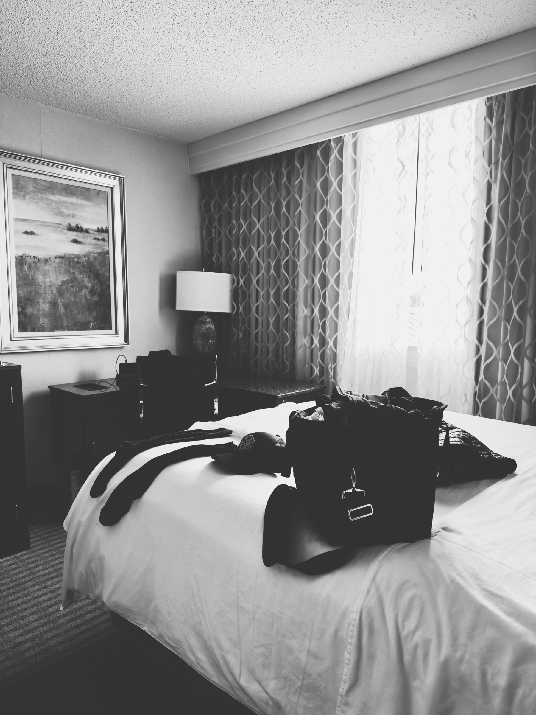 Auto-generated description: A neatly made hotel bed is adorned with bags and clothing, set beside a window with patterned curtains and a framed piece of art on the wall.