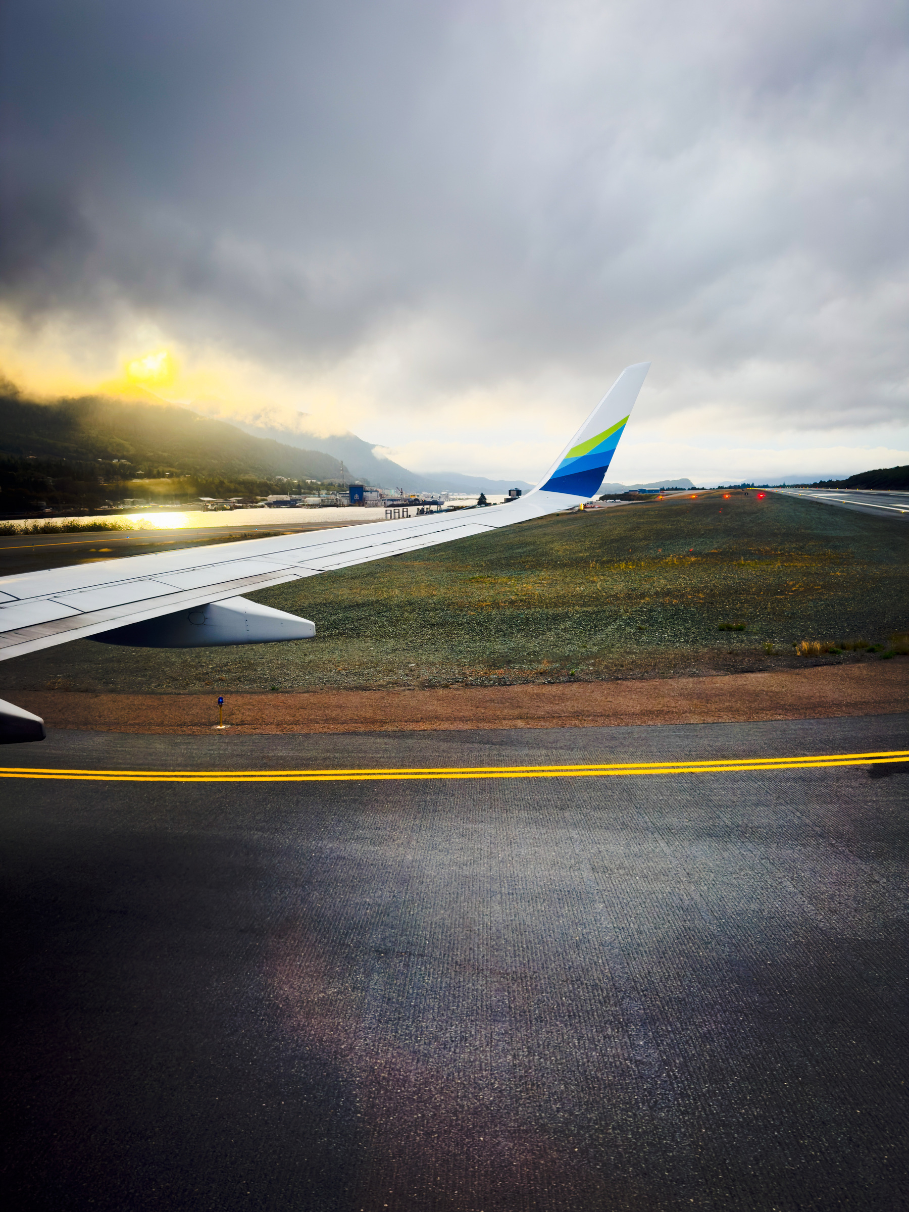 Auto-generated description: An airplane wing is visible from the window seat as the plane taxis on a runway, with dramatic clouds and a sunset in the background.