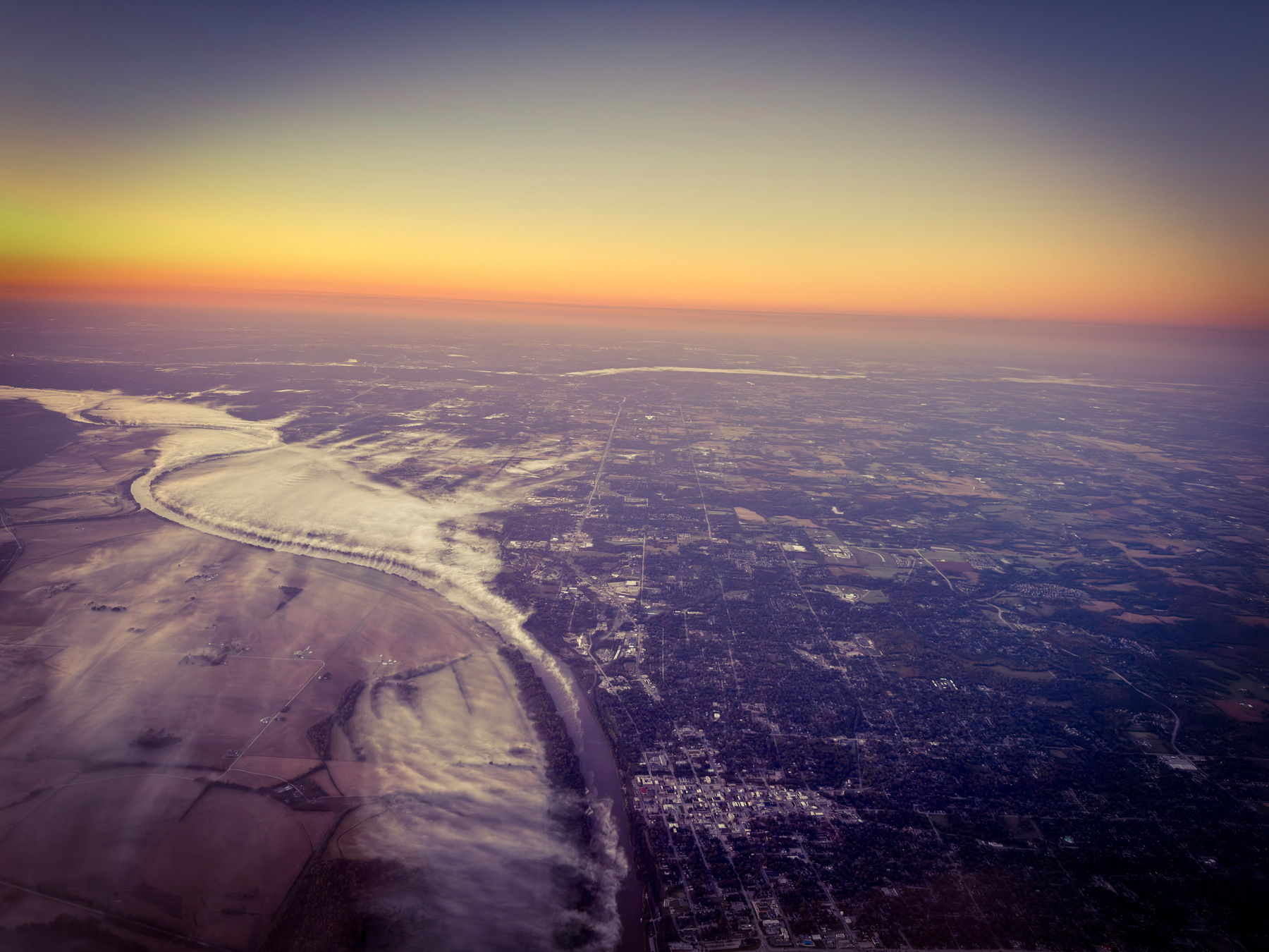 Auto-generated description: An aerial view showcases a river winding through a landscape with farmlands and urban areas beneath a colorful sunset sky.
