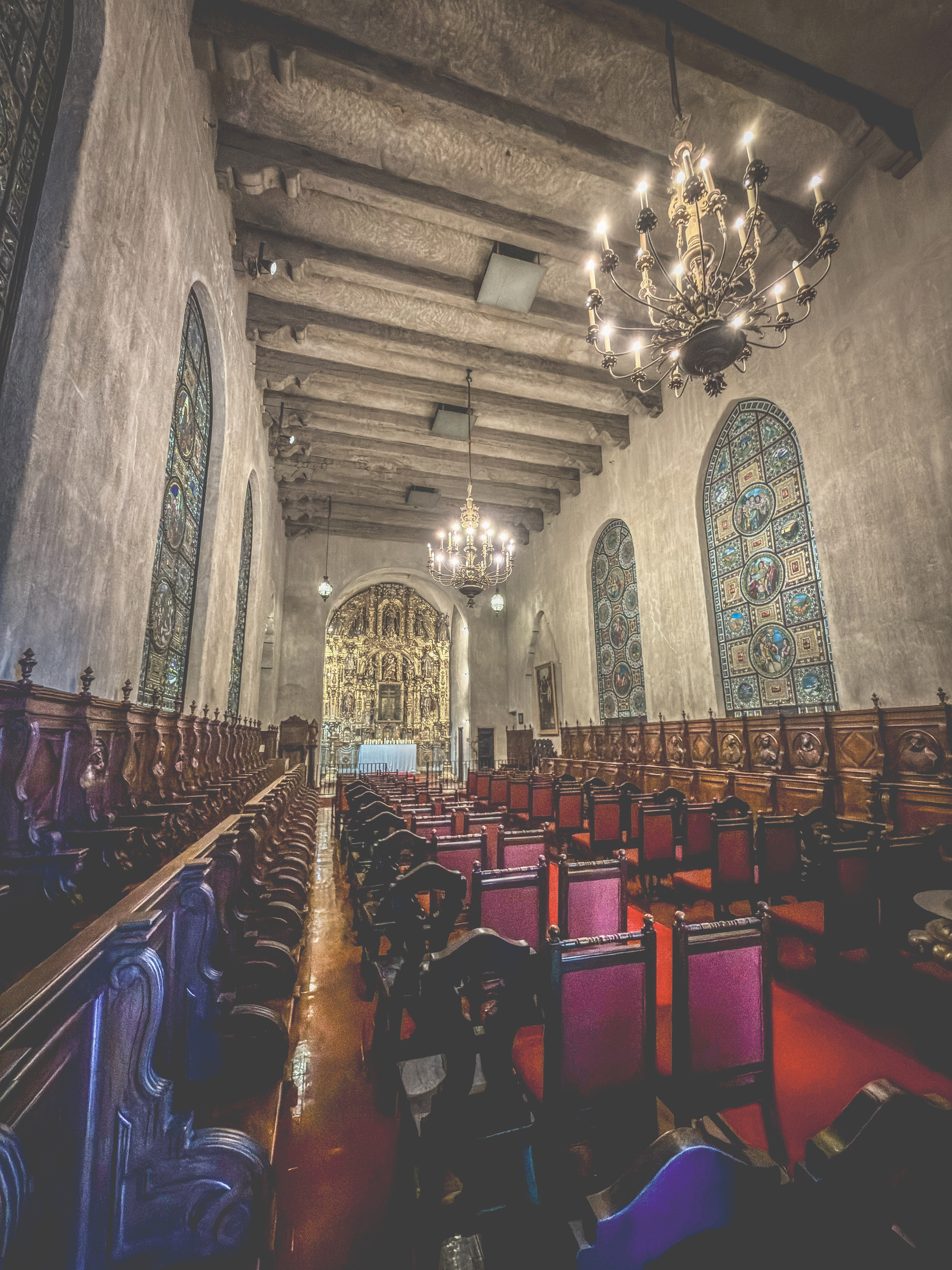 Auto-generated description: A historic chapel with ornate chandeliers, stained glass windows, and rows of wooden pews leads to an intricately designed altar.