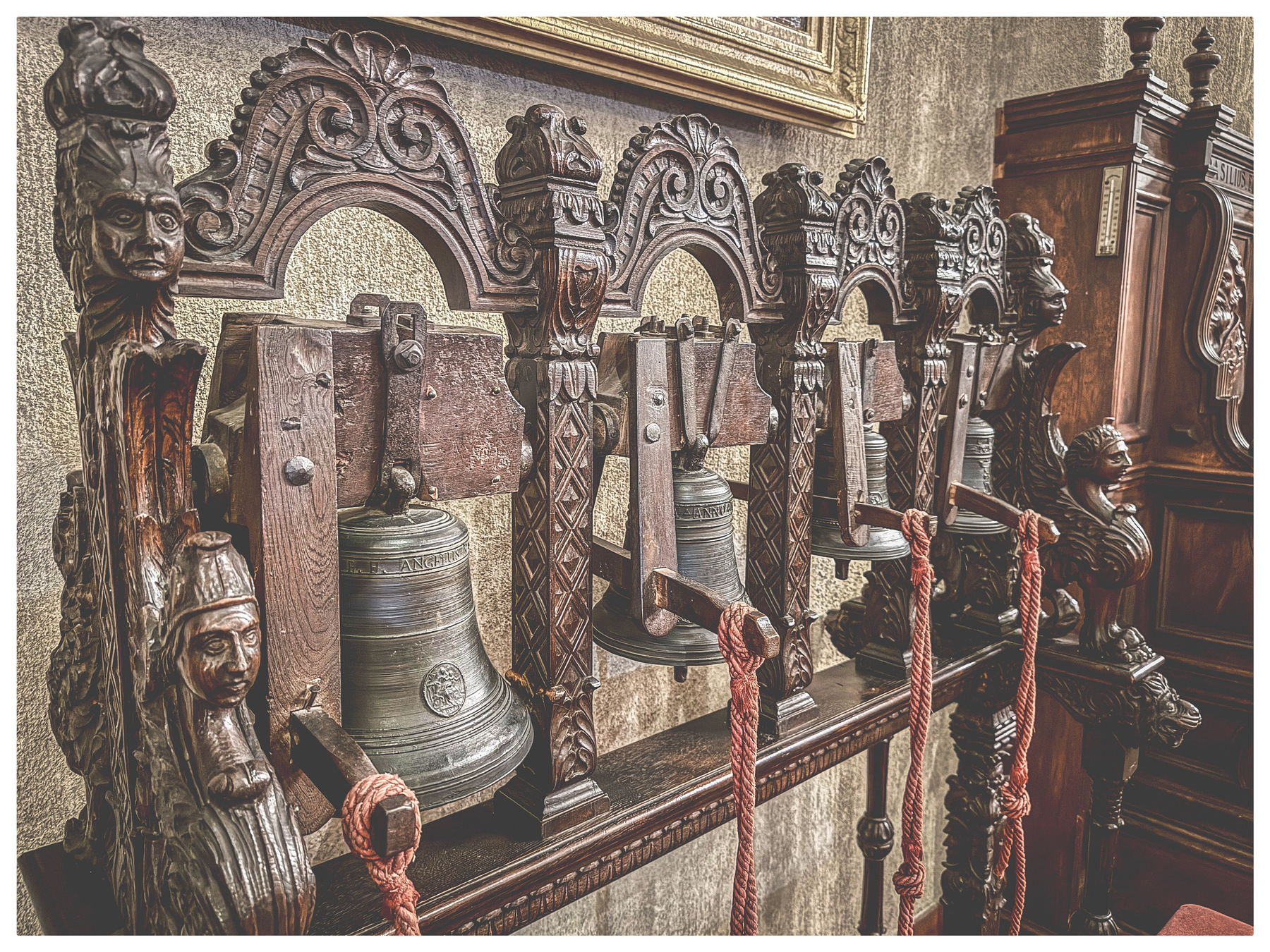 Auto-generated description: There is an ornate, antique wooden rack holding several old metal service bells with intricate carvings and ropes.