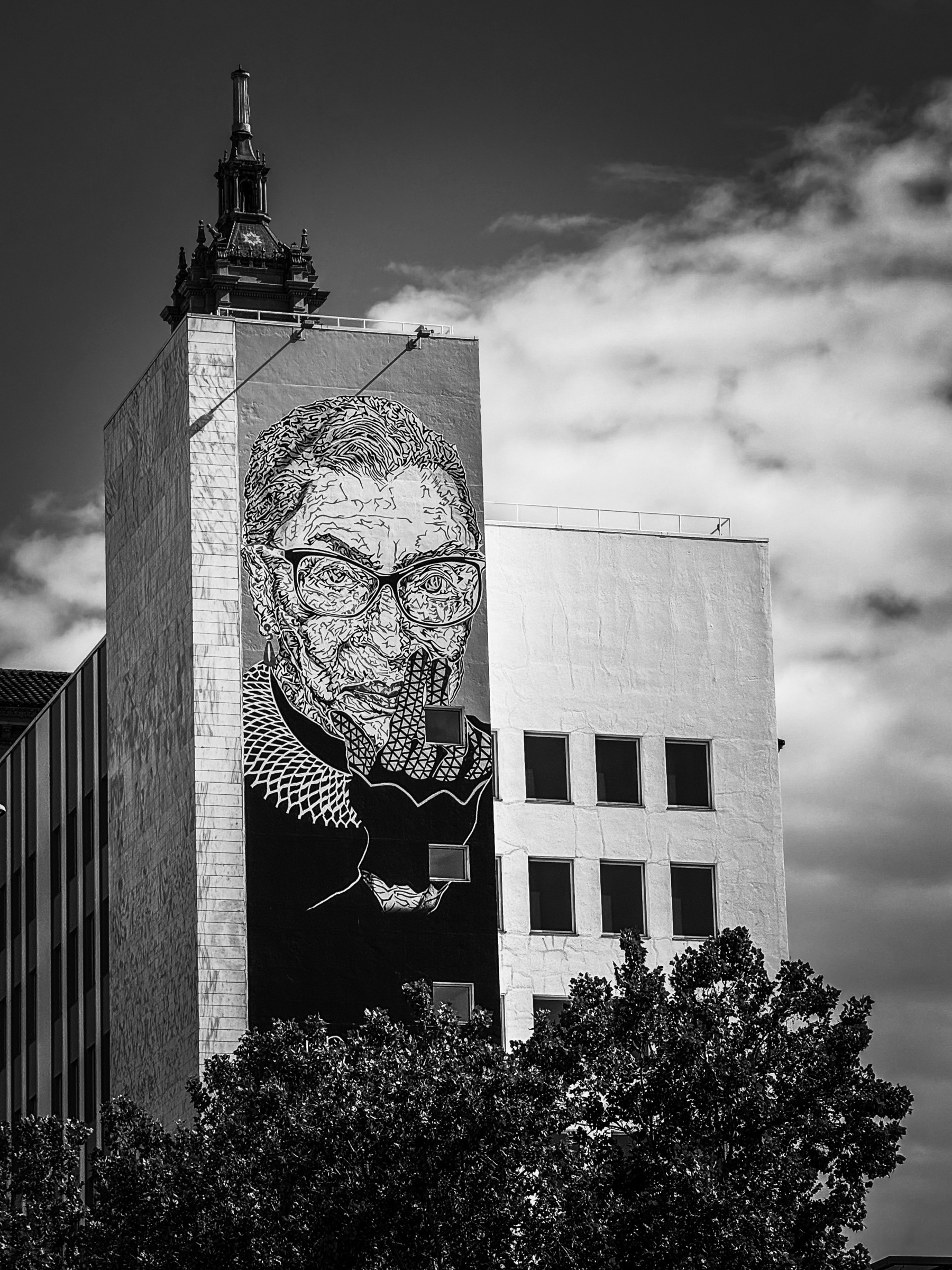 Auto-generated description: A large mural featuring a detailed, stylized portrait of a person adorns the side of a multi-story building, with trees in the foreground.