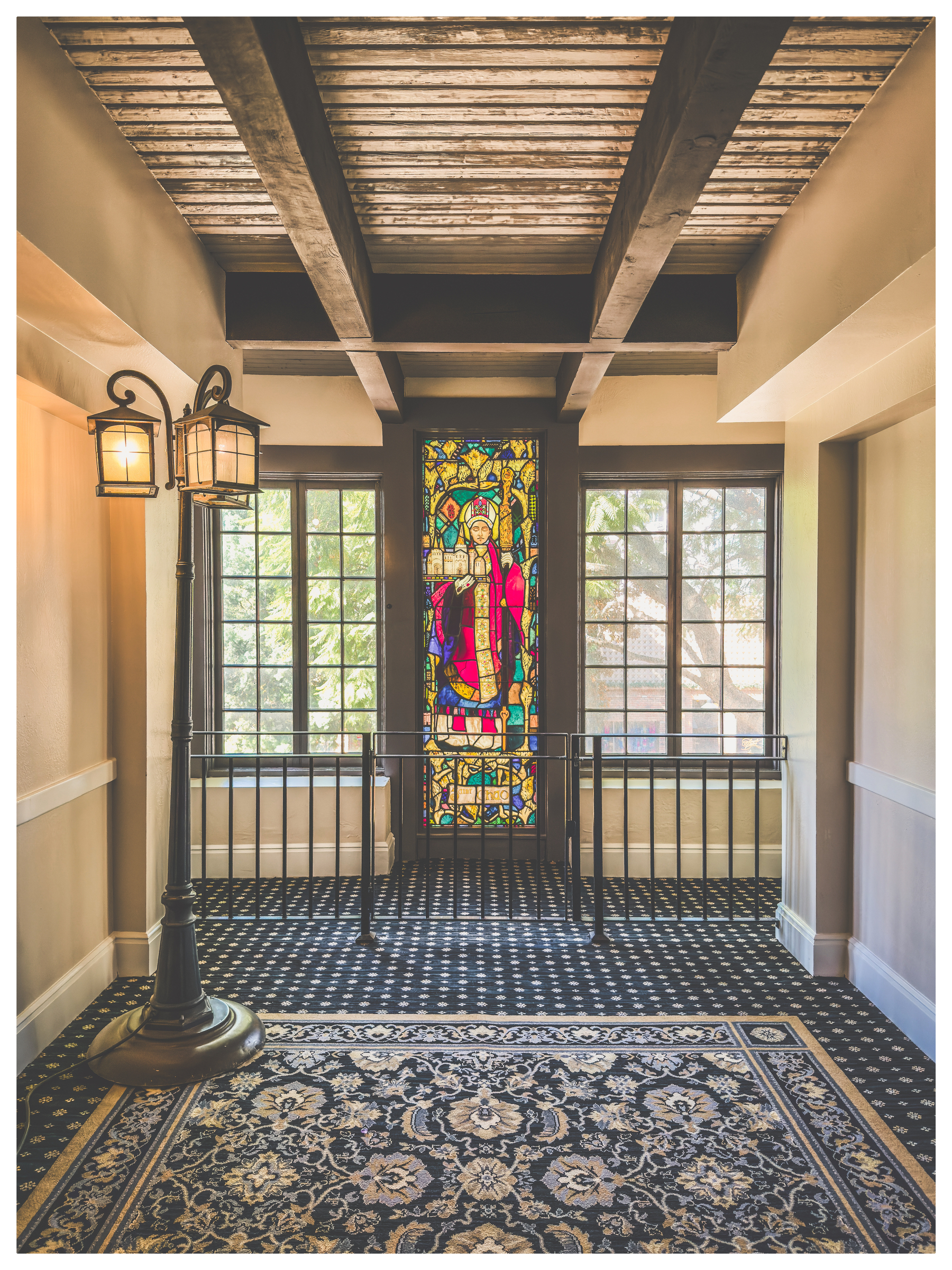 Auto-generated description: A hallway features a stained glass window with a colorful design, flanked by two windows and illuminated by a vintage-style floor lamp.
