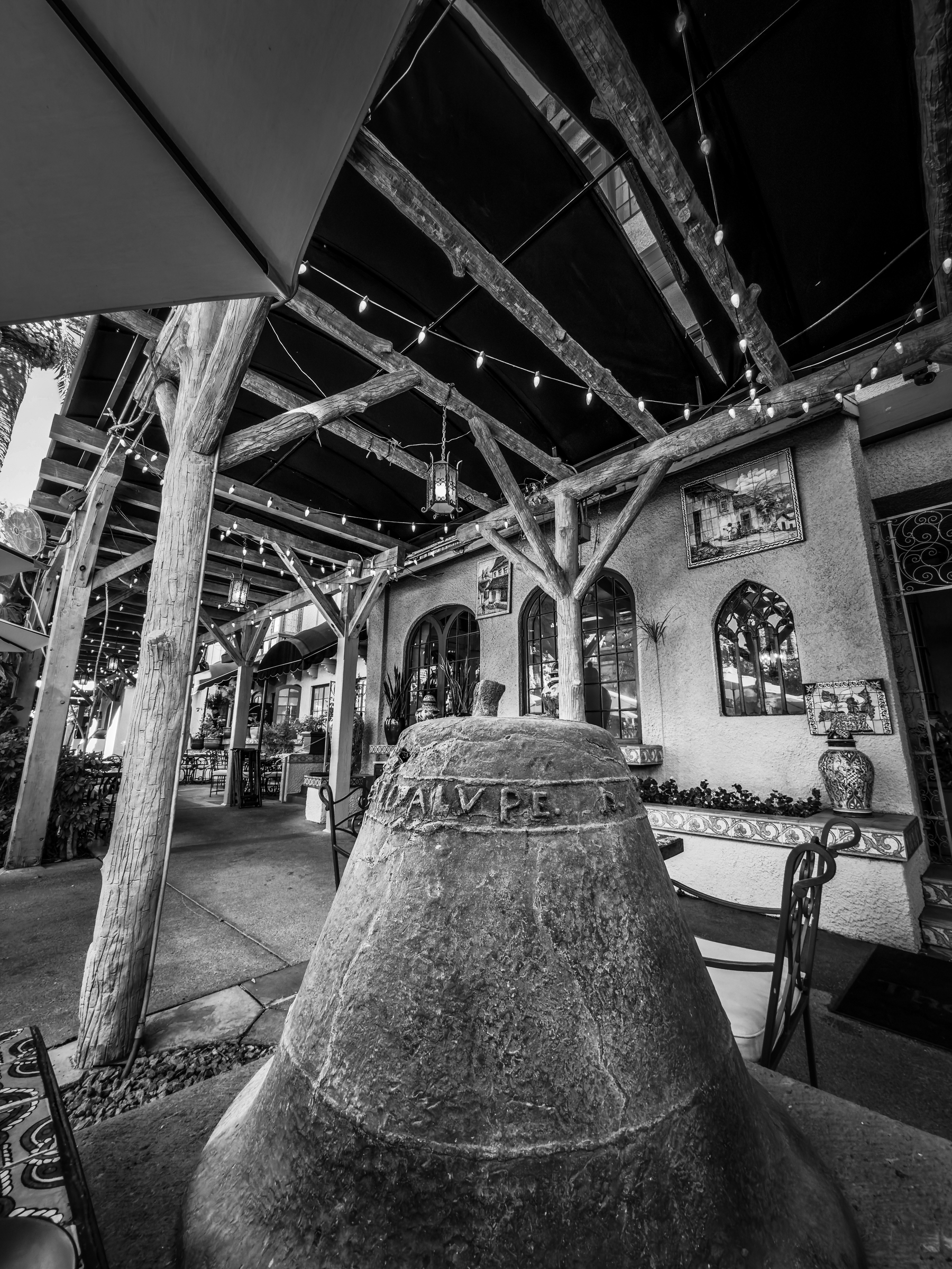 Auto-generated description: A rustic outdoor courtyard features a large historic bell in the foreground, surrounded by wooden beams and adorned with string lights.