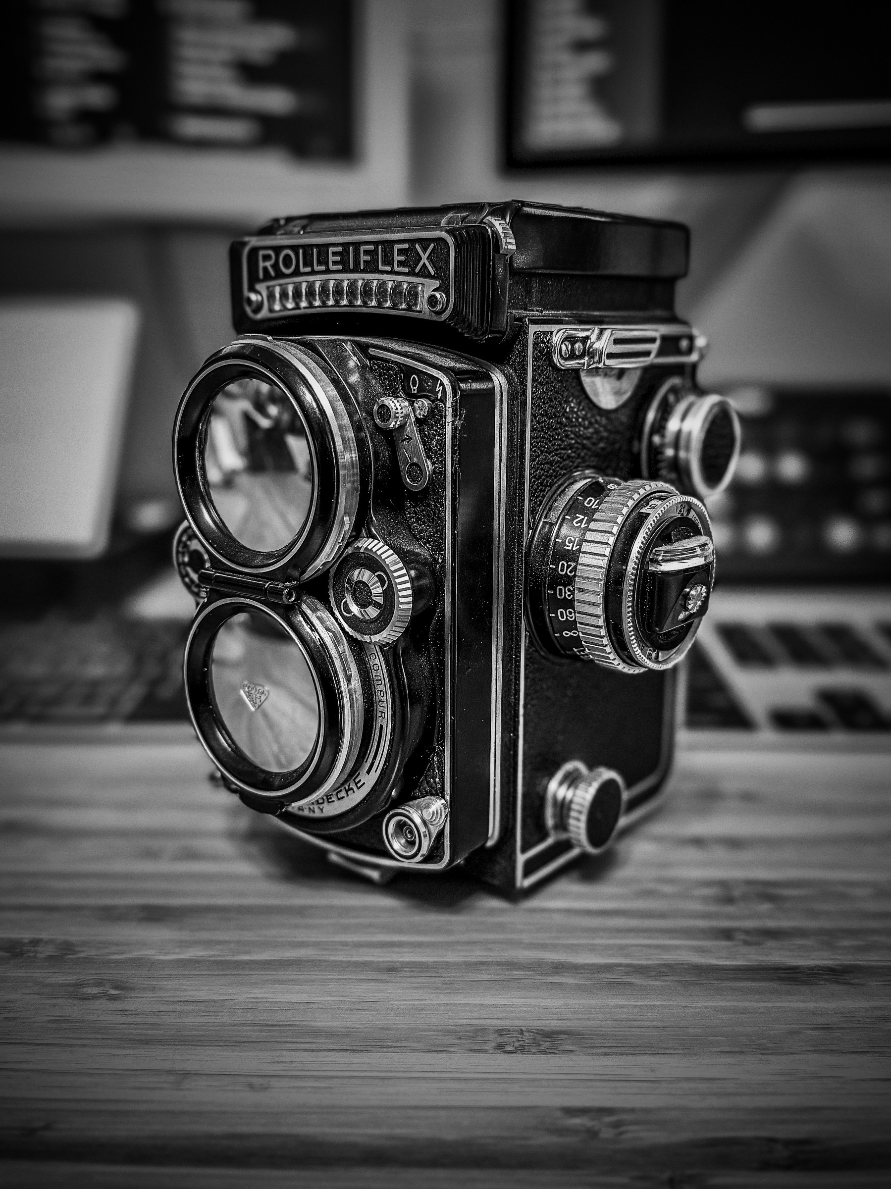 Auto-generated description: A vintage Rolleiflex twin-lens reflex camera is displayed on a wooden surface, with a blurred background.