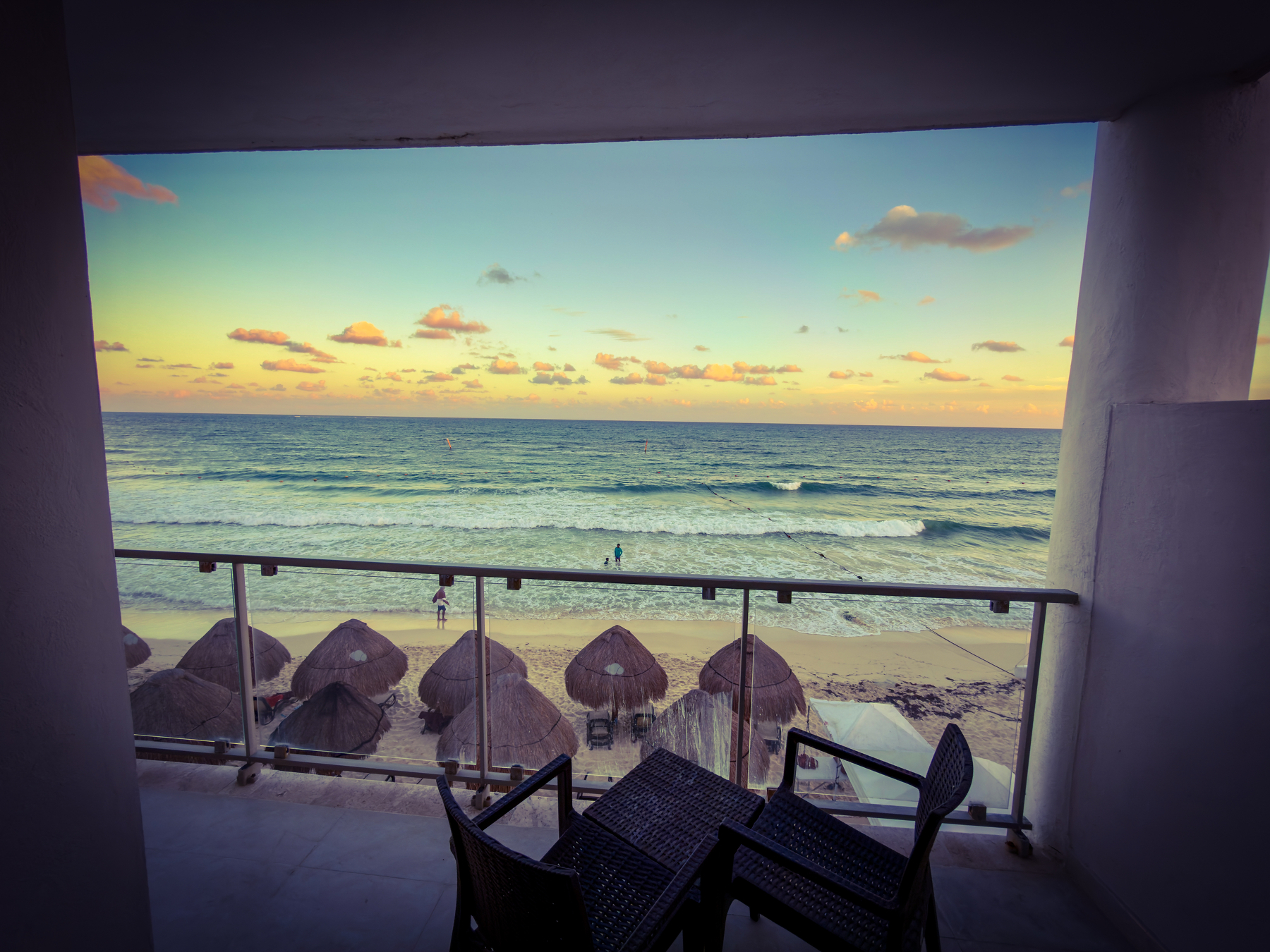 Auto-generated description: A balcony overlooking a beach with straw umbrellas and a colorful sunset over the ocean.