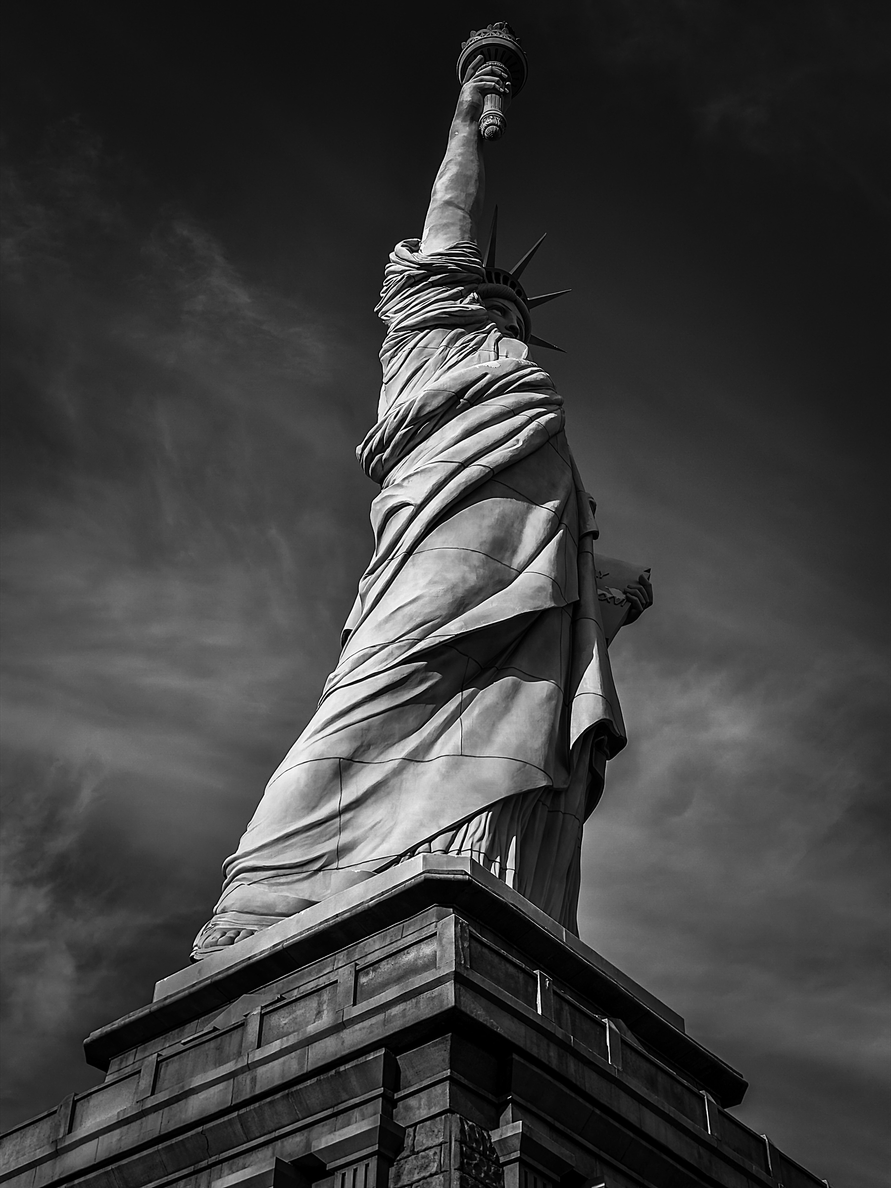 Auto-generated description: A dramatic black and white photograph of the Statue of Liberty viewed from the back, highlighting its towering presence against a cloudy sky.
