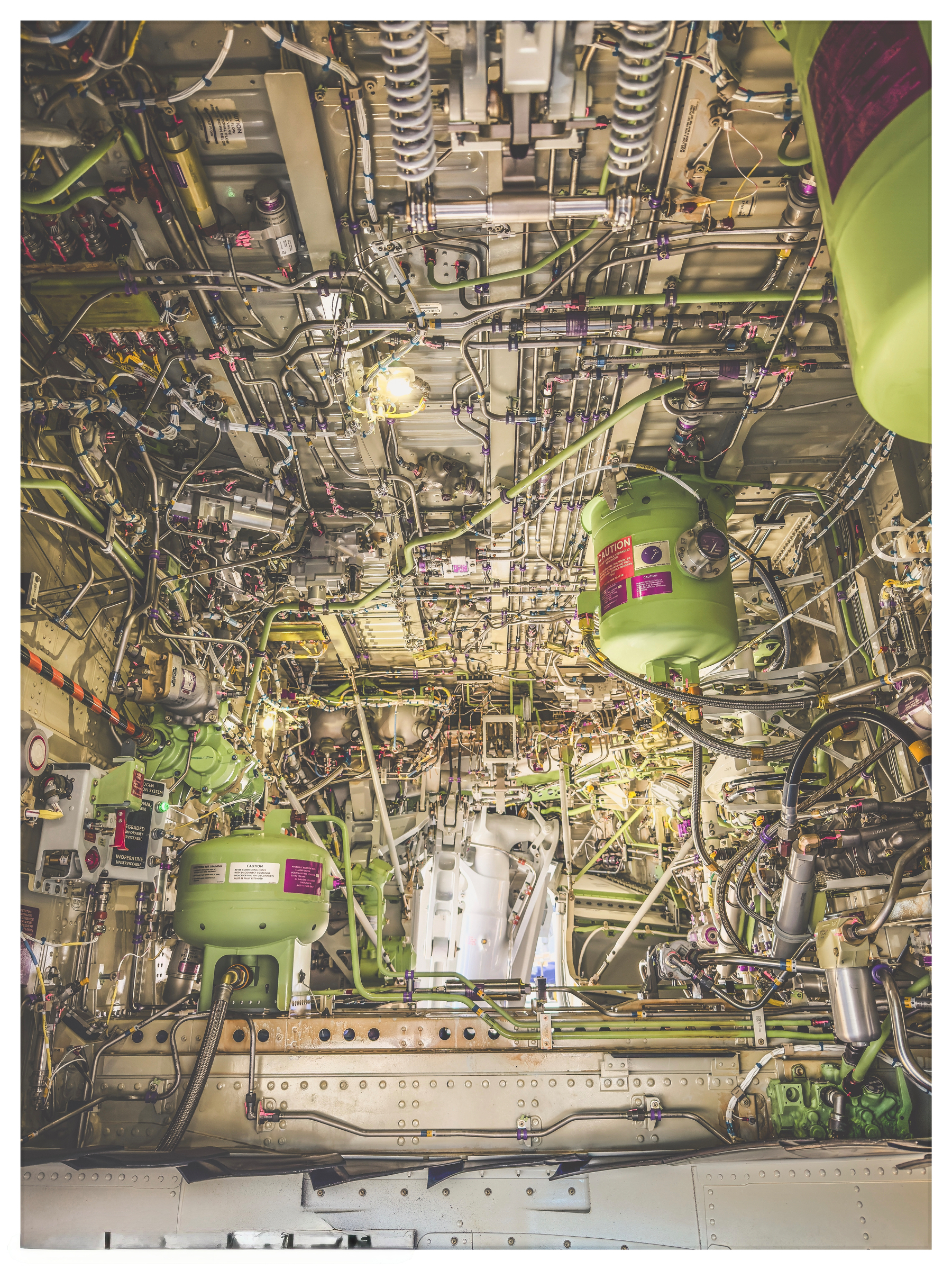 Auto-generated description: A complex network of cables, pipes, and machinery parts with green cylindrical components is visible inside an aircraft or industrial compartment.