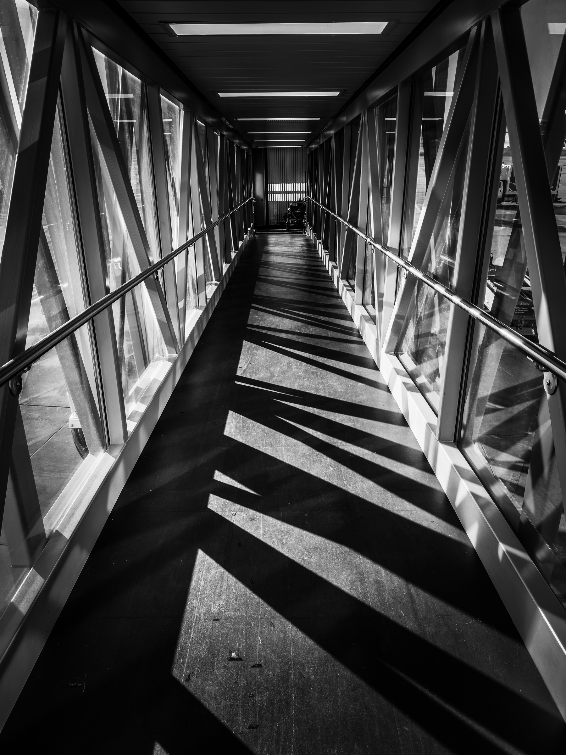 Auto-generated description: A long, empty glass corridor with metal beams casts geometric shadows on the floor.